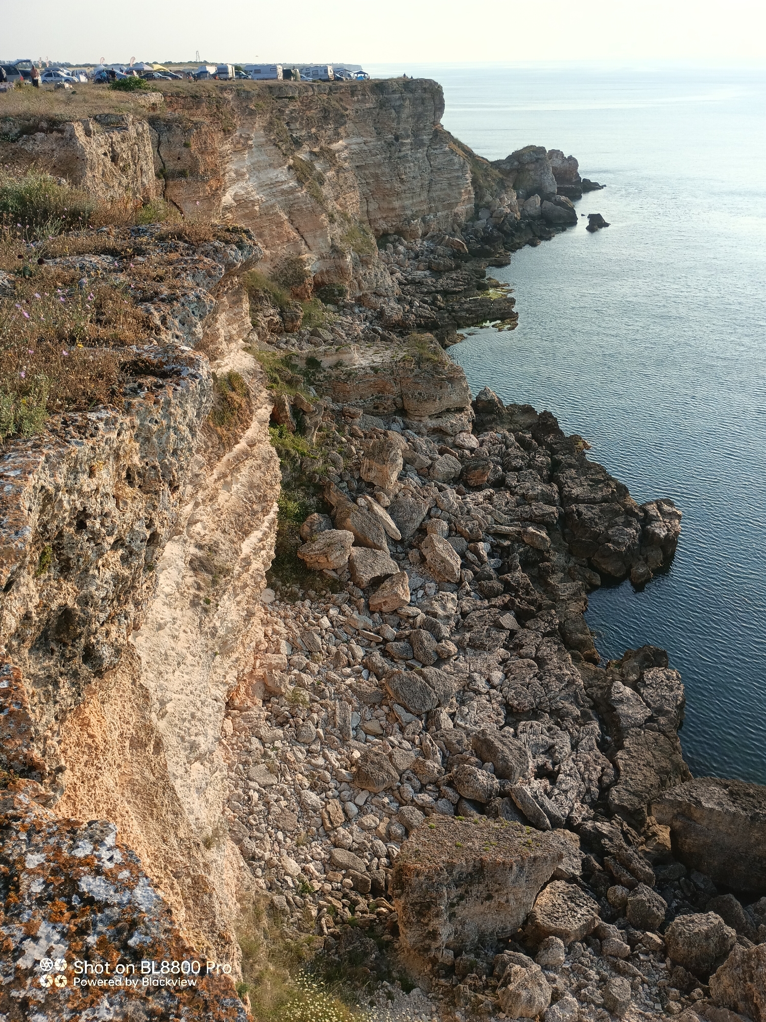 The image depicts a rugged cliffside overlooking a calm sea. The cliffs are composed of layered sedimentary rock, with visible strata and some areas of erosion. Sparse vegetation, including dry grasses and a few small purple flowers, grows on the cliff tops and ledges. Below the cliffs, a jumble of large rocks and boulders forms a rocky shoreline. The sea is a deep blue with gentle ripples. In the distance, the horizon is clear, with a pale sky. On the very top of the cliff in the background, several recreational vehicles, tents, and cars are parked, suggesting a campsite or a public gathering area. The overall scene is natural and serene, with human presence limited to the distant vehicles. The image captures the natural beauty of the coastline in Kamen bryag, Bulgaria, known for its dramatic cliffs and clear waters.