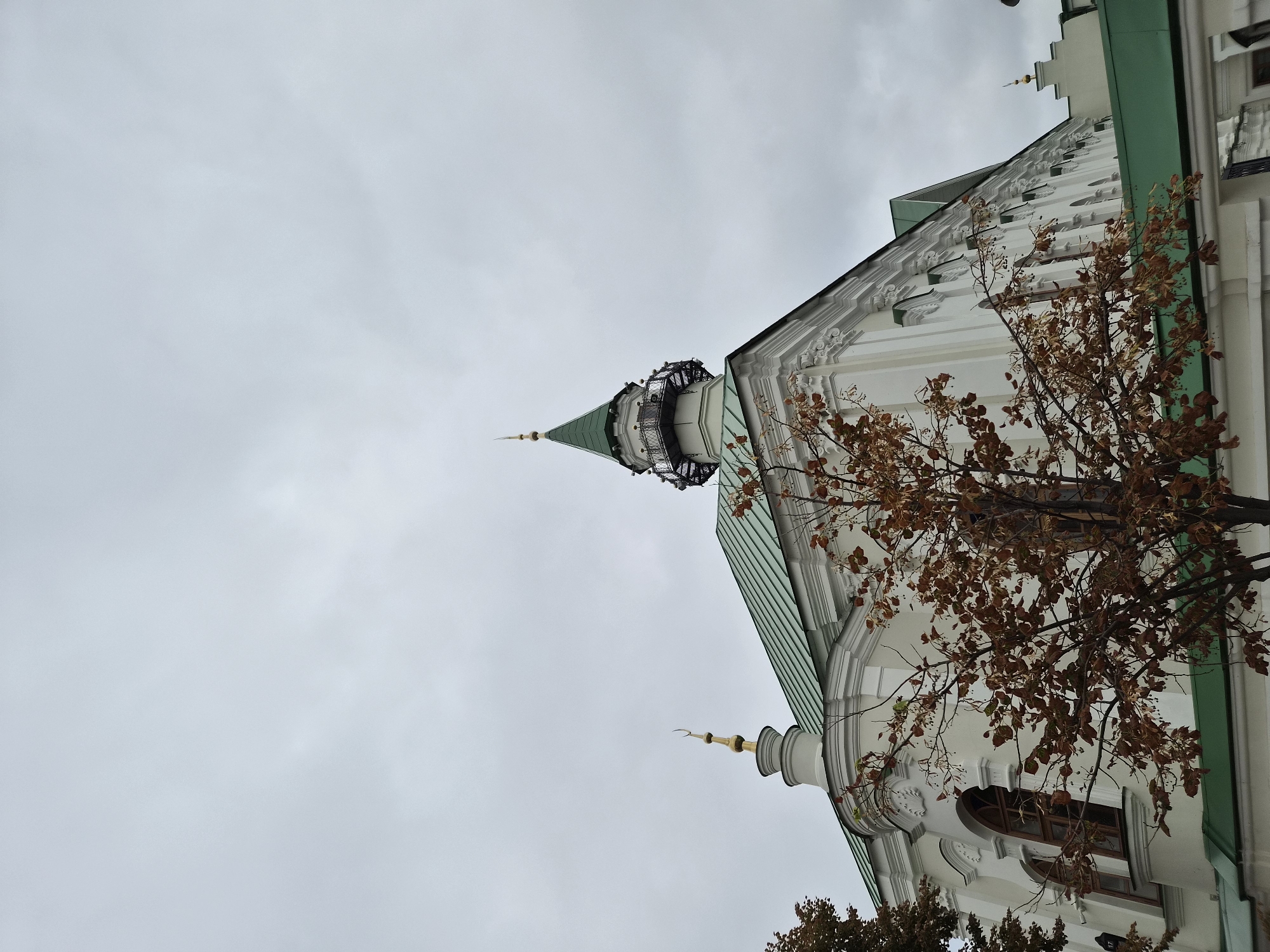 The image shows a side view of the Kul Sharif Mosque in Kazan, Russia, on a cloudy day. The mosque features a white facade with ornate detailing, a green tiled roof, and a tall, slender minaret topped with a green spire and a golden crescent moon. A tree with brown, autumnal leaves is in the foreground, partially obscuring the mosque. The sky is overcast and gray. There are no people visible in the image.