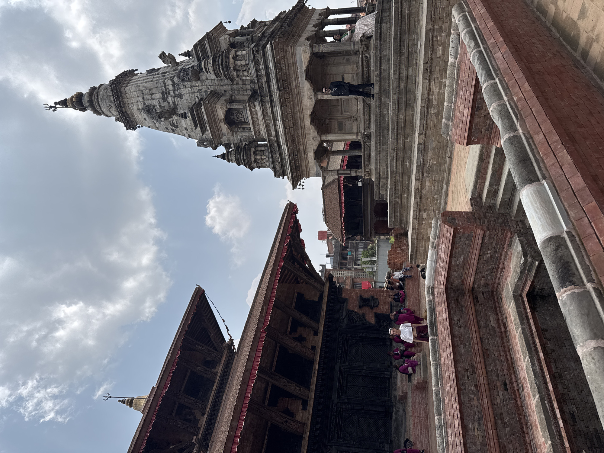 The image shows a view of Bhaktapur Durbar Square in Nepal, featuring a traditional multi-tiered pagoda-style building with intricate wooden carvings and a brick structure. In the background stands a taller, stone temple with a spire. A man with dark clothes is standing on top of the multi-tired building. Monks are sitting on the floor in front of the building with wooden carvings. The sky is cloudy.