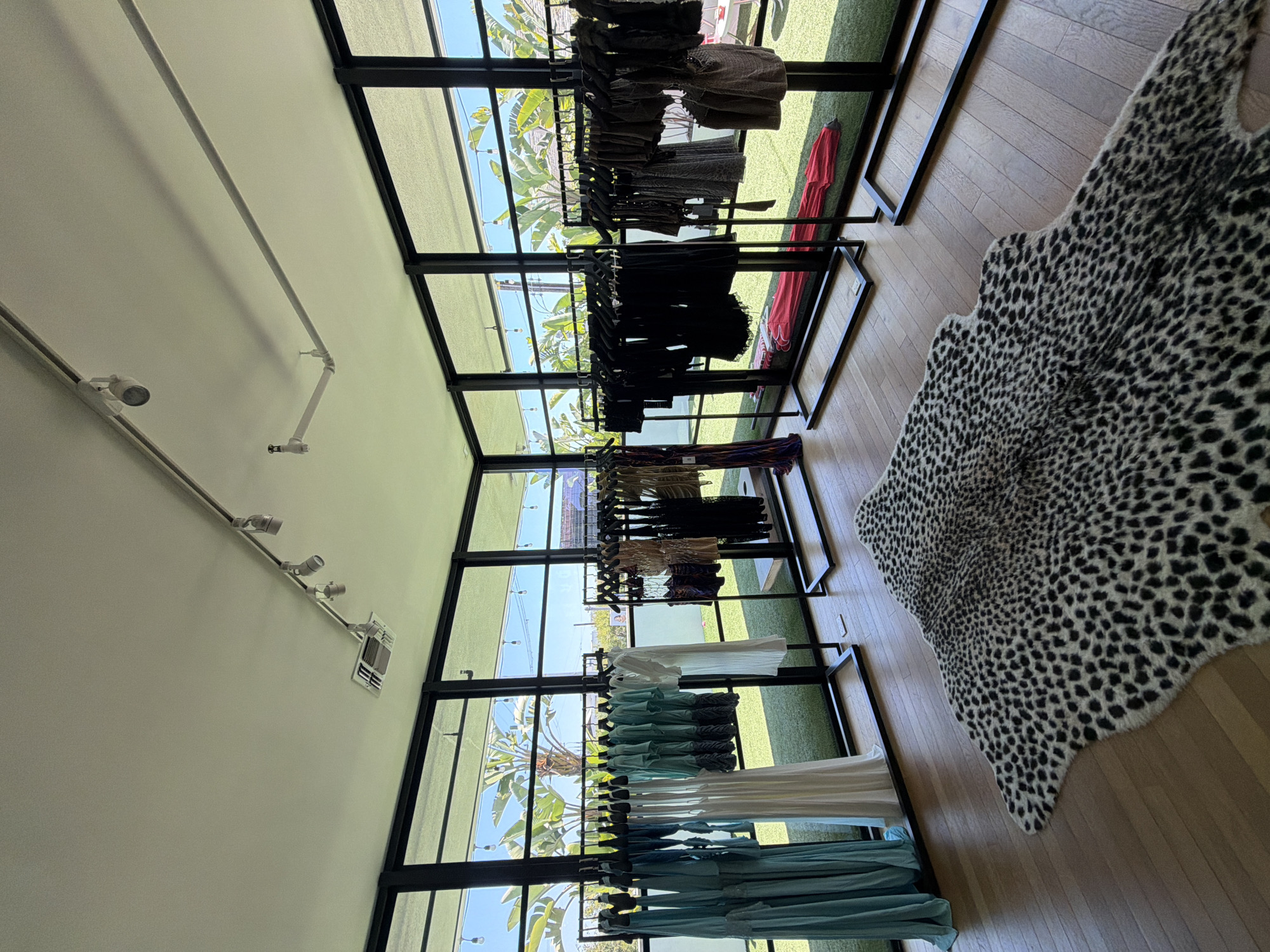 The image shows the interior of a clothing boutique in West Hollywood, California, United States. The boutique features a series of black-framed, floor-to-ceiling windows providing ample natural light and views of palm trees and greenery outside. Clothing racks are arranged along the length of the windows, displaying apparel in various colors and styles, from dark tones to lighter shades of blue and white. The floor is made of light-colored wooden planks, partially covered by a faux animal hide rug with a black and white spotted pattern. The ceiling is white, with a track lighting system running along its length. The scene is well-lit and appears to be taken during daylight hours.