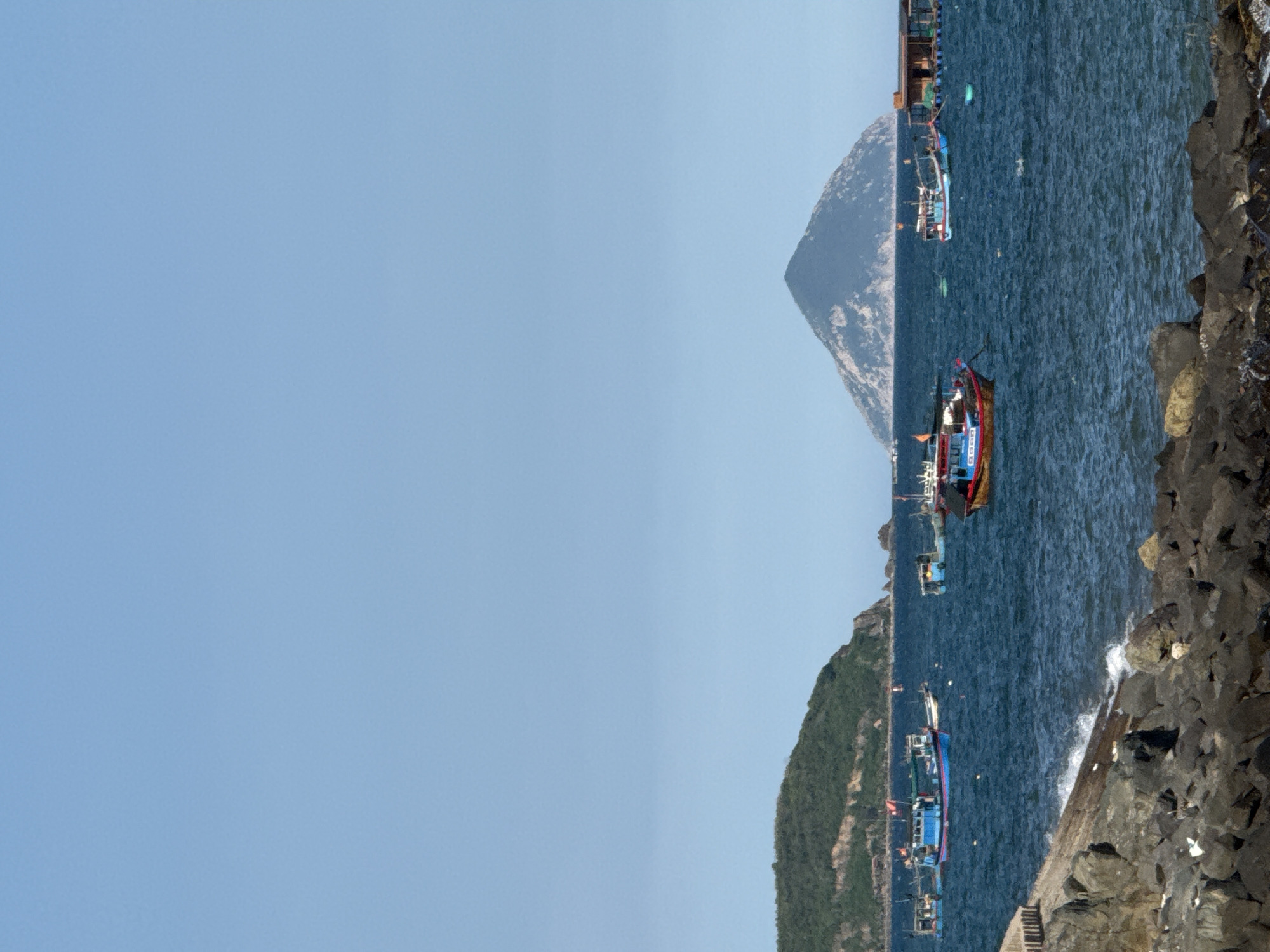 The image depicts a coastal scene with several fishing boats on the water. In the foreground, a rocky shore is visible, with waves gently lapping at the rocks. Several boats of varying sizes and colors are scattered across the blue water. Some boats are closer to the shore, while others are further out. In the background, a lush green island dominates the left side of the frame, and a distinct conical mountain rises from the sea on the right. The sky is clear and blue, indicating a sunny day. The scene suggests a typical day for a fishing community. This location is identified as being in Bac Nha Trang Ward, Vietnam, a region known for its coastal beauty and fishing activities.