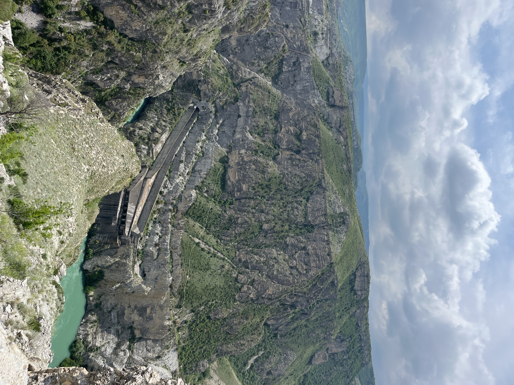 The image depicts a landscape with a notable hydroelectric dam feature. Located in Chirkey, Russia, the scene presents a deep gorge or canyon carved by a river flowing through it, exhibiting a turquoise color. The dam structure is visible, constructed of gray concrete and extending across the gorge. The steep canyon walls show rocky terrain with sparse vegetation. The sky above is partly cloudy, suggesting fair weather. The overall perspective is from a high vantage point looking downward. There are no discernible people in the image.