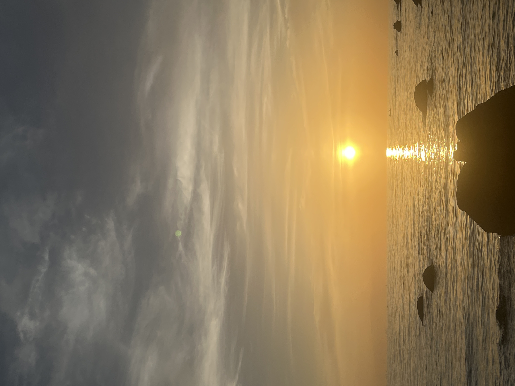 The image depicts a serene sunset over a calm body of water, likely the sea, with several dark, rounded rocks partially submerged in the foreground and scattered across the surface. The sun is a bright, radiant orb near the horizon, casting a golden glow and a shimmering reflection on the water. The sky transitions from a dark, cloudy grey at the top to a vibrant orange and yellow as it approaches the sun. In the distance, a faint, hazy silhouette of landmasses can be seen. The overall impression is one of tranquility and natural beauty, possibly a coastal scene in the Ko Pha-ngan District of Thailand, known for its picturesque beaches and sunsets.