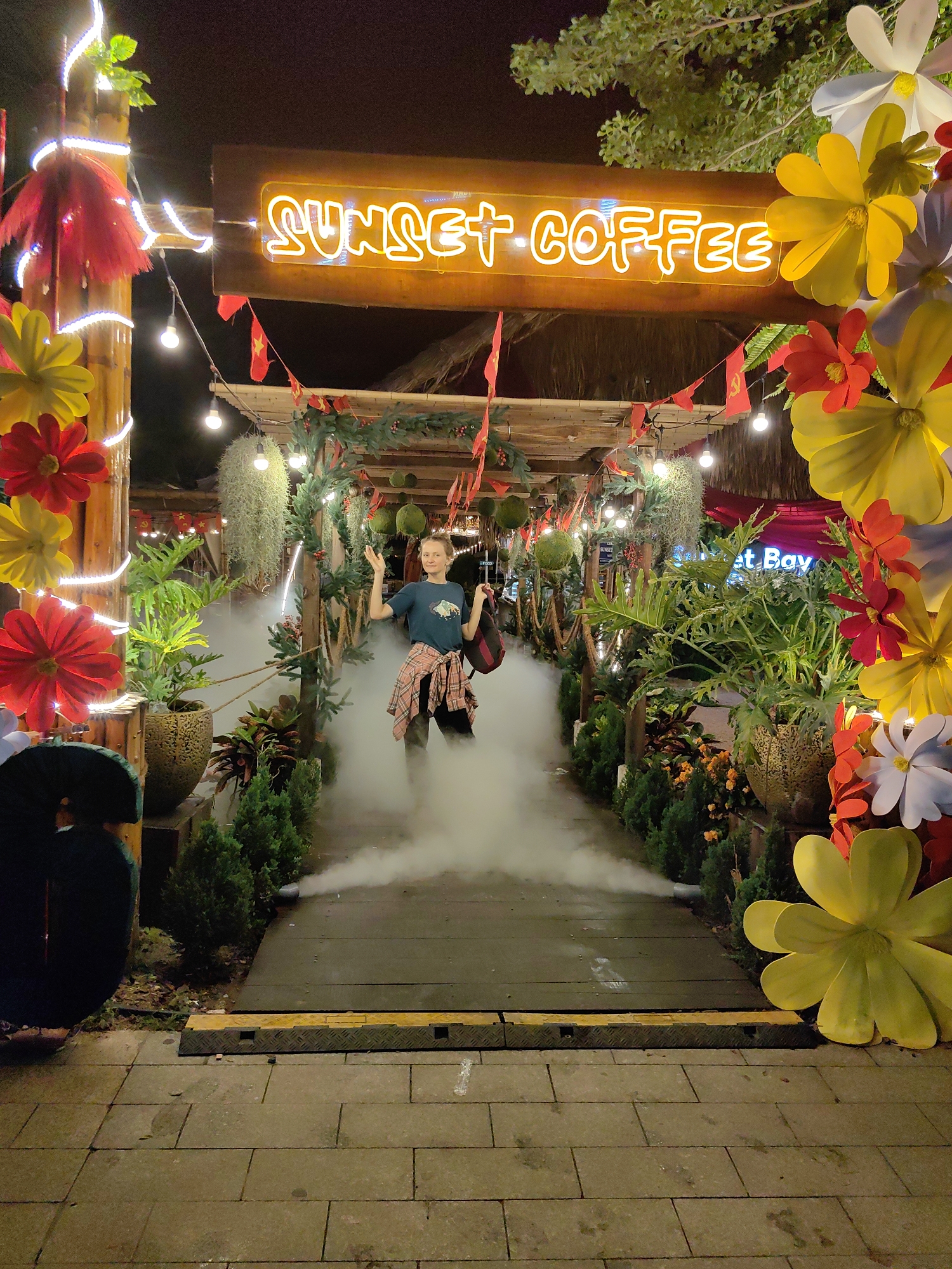 An eye-level, full shot captures a woman posing playfully at the illuminated entrance of a coffee shop named "Sunset Coffee" at night. The scene is richly decorated with a festive and tropical theme.
The main subject, a woman with light-colored hair, stands centrally on a wooden pathway, enveloped by artificial fog or smoke emanating from ground-level sources. She wears a dark blue t-shirt and has a red and black plaid shirt tied around her waist, looking towards the viewer with a slight smile and raised hands. A dark shoulder bag hangs from her left shoulder.
The entrance is framed by bamboo poles wrapped with string lights and adorned with large artificial flowers in red, yellow, and white. Overhead, a prominent orange neon sign glows with the words "SUNSET COFFEE" in a stylized font. The archway and surrounding structures are draped with lush greenery, including potted plants, hanging tillandsias, and decorative moss balls. Small red flags, some featuring a yellow star reminiscent of the Vietnamese flag, are strung across the top, providing a subtle location cue alongside the provided context of Bai Chay Ward, Vietnam.
String lights illuminate the dark night sky. In the background, further lights and structures are visible, with a partially obscured sign reading "et Bay" on the right. The ground leading to the entrance is paved with stone tiles, and a dark, textured ramp-like structure marks the transition to the wooden path. The overall ambiance is inviting, whimsical, and designed to attract evening visitors to the establishment.