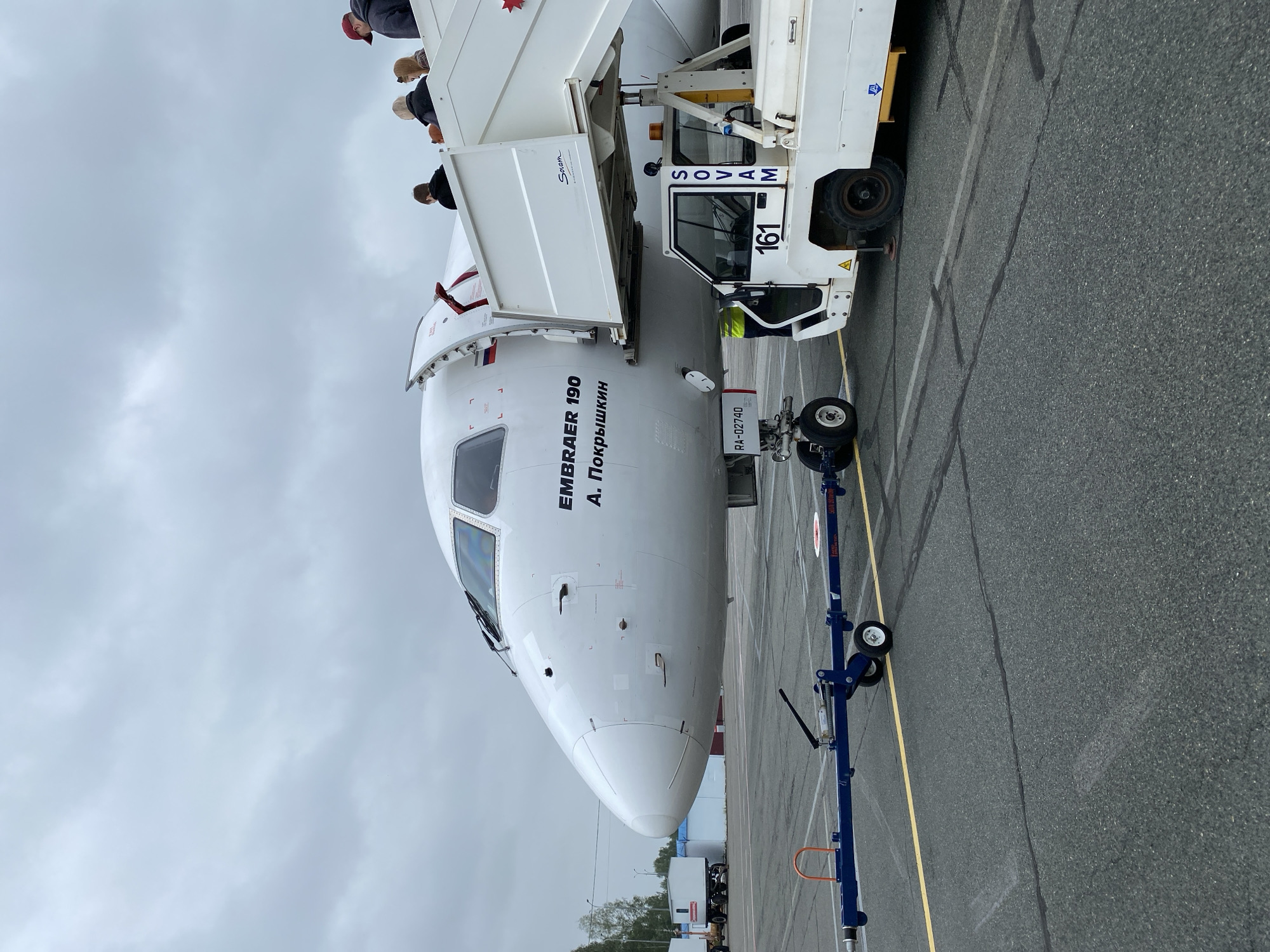 An Embraer 190 aircraft, identified by the text "EMBRAER 190 А. Покрышкин" and registration "RA-02740", is parked on an asphalt tarmac under a cloudy sky. Passengers are boarding the aircraft via a mobile staircase. A white ground support vehicle with the number "161" and the word "SOVAM" is positioned near the aircraft's nose. A blue tow bar is attached to the aircraft's front landing gear. In the background, there are small white trailers and trees. The scene depicts typical airport operations.