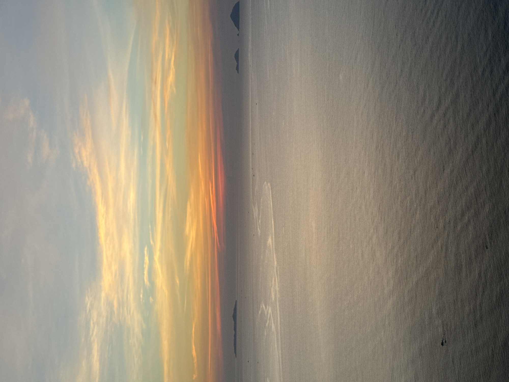 The image shows a serene seascape at sunset in Nha Trang Ward, Vietnam. The sky displays a gradient of colors, transitioning from a soft blue at the top to hues of orange and pink near the horizon, indicative of the setting sun. Wispy clouds are scattered across the sky. In the distance, several small islands or landmasses are visible on the horizon. The sea is calm, with subtle ripples on its surface, reflecting the colors of the sky. A faint, winding trail, possibly from a boat or currents, is visible on the water in the mid-ground. In the foreground, a small boat with two figures is present on the water, heading towards the left side of the frame. The overall scene is tranquil and expansive, capturing the natural beauty of the coastal environment.