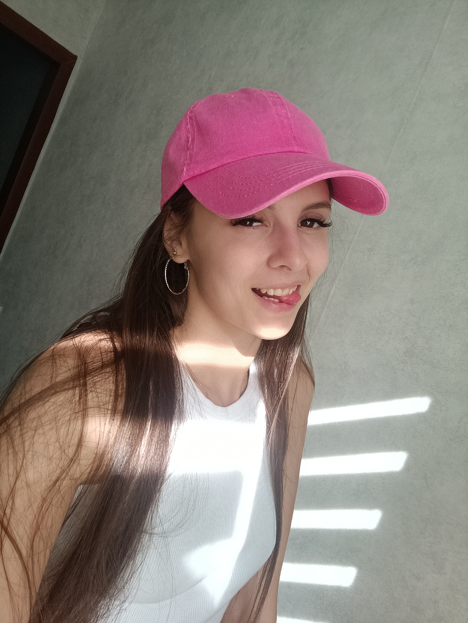 A young woman is standing indoors. She is wearing a pink baseball cap, a white tank top, and gold hoop earrings. She is sticking out her tongue slightly and smiling. Her hair is long and brown. Light streams in through window blinds, casting horizontal shadows across the wall behind her. To the left is a dark-framed window or door. The location is Unknown, Unknown.