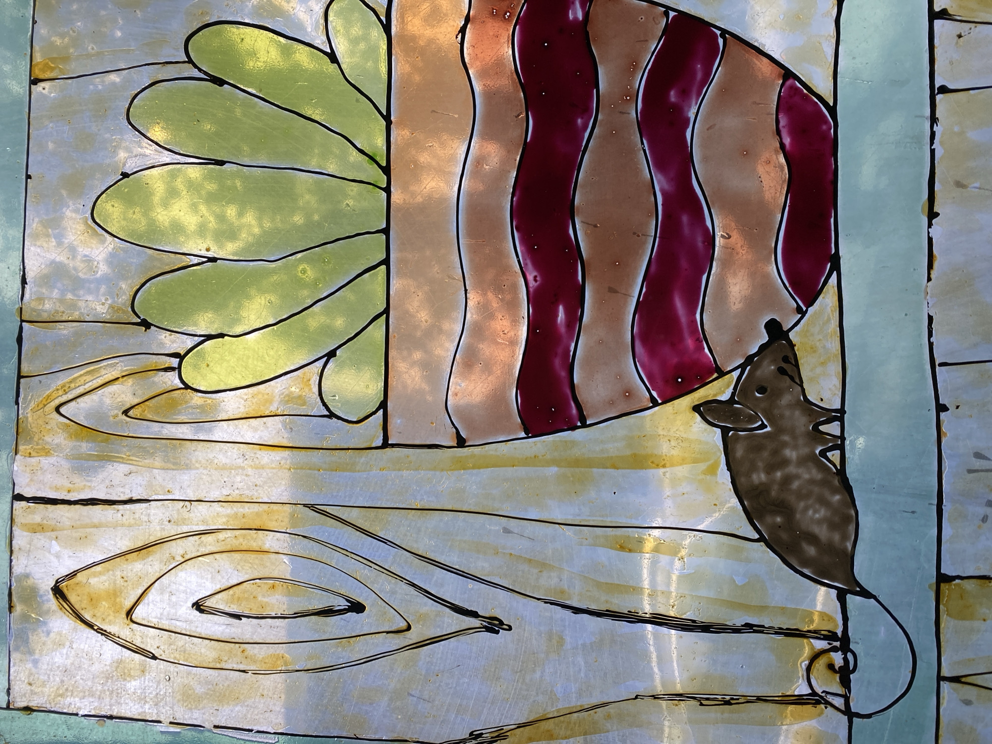 The image displays a close-up of stained glass artwork. The artwork depicts a potted plant with large, light green leaves, outlined in black. The pot itself has a striped design in shades of copper and dark red. To the left of the pot, a small brown mouse with a long, curly tail is positioned on a light blue surface. The background of the stained glass features abstract swirling patterns in yellow and white, resembling wood grain. This artwork is not located in the city of Poltava, Ukraine; it is a general description of the artwork itself.