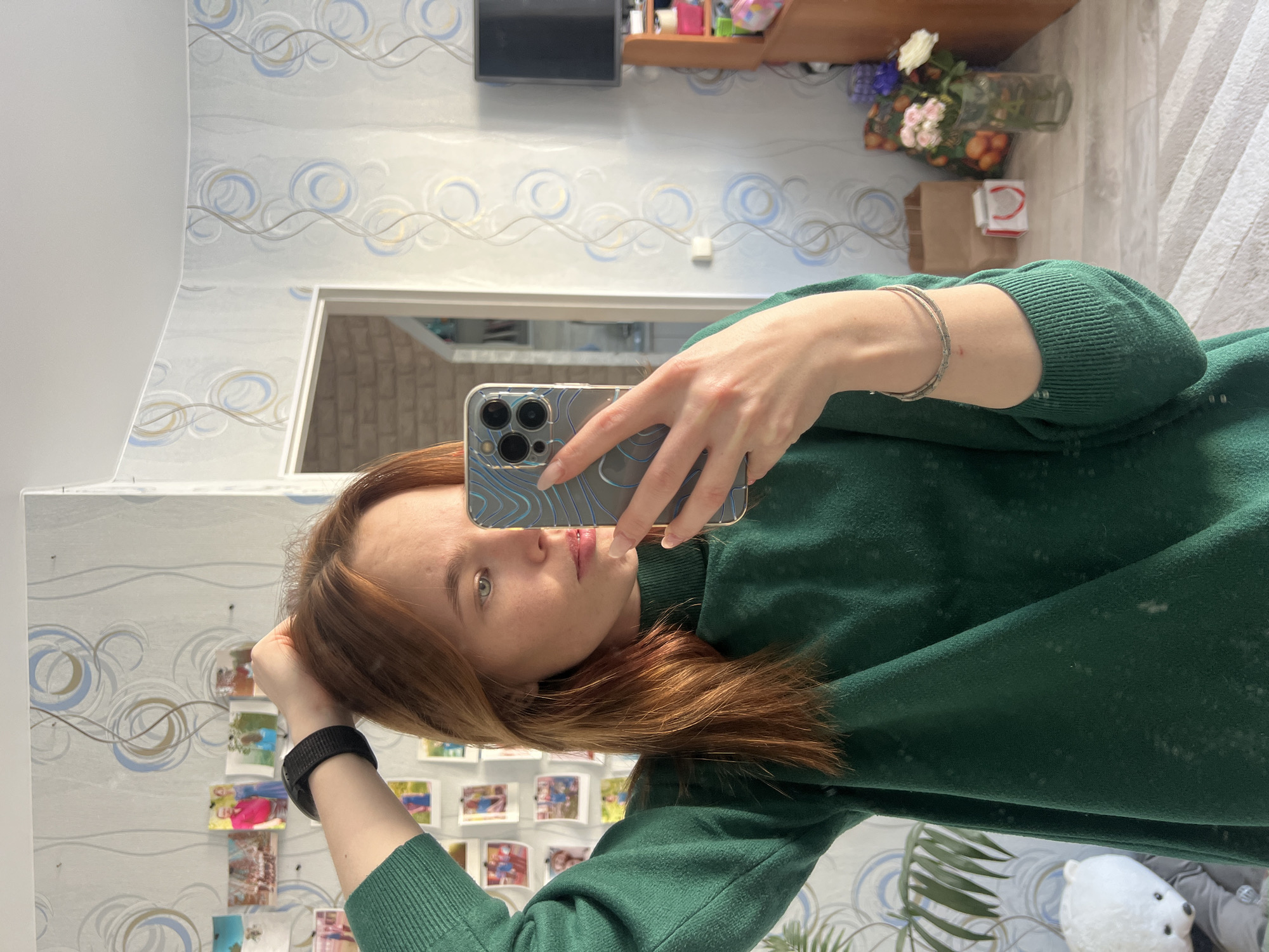 A person takes a selfie in a room with wallpaper featuring blue and gold swirls. The person is fair-skinned with auburn hair and is wearing a green sweatshirt. They hold a smartphone with a clear case in their right hand. A silver bracelet is visible on their right wrist, and a black smartwatch is on their left. Behind the person, a mirror reflects photos attached to the wall. A doorway is visible in the background, leading to another room. A TV, shelves with objects, flowers in a vase, and fruits are placed on top of a wooden surface behind the person's head. The location appears to be an interior space. No specific landmarks indicate the city of Chaikovsky.