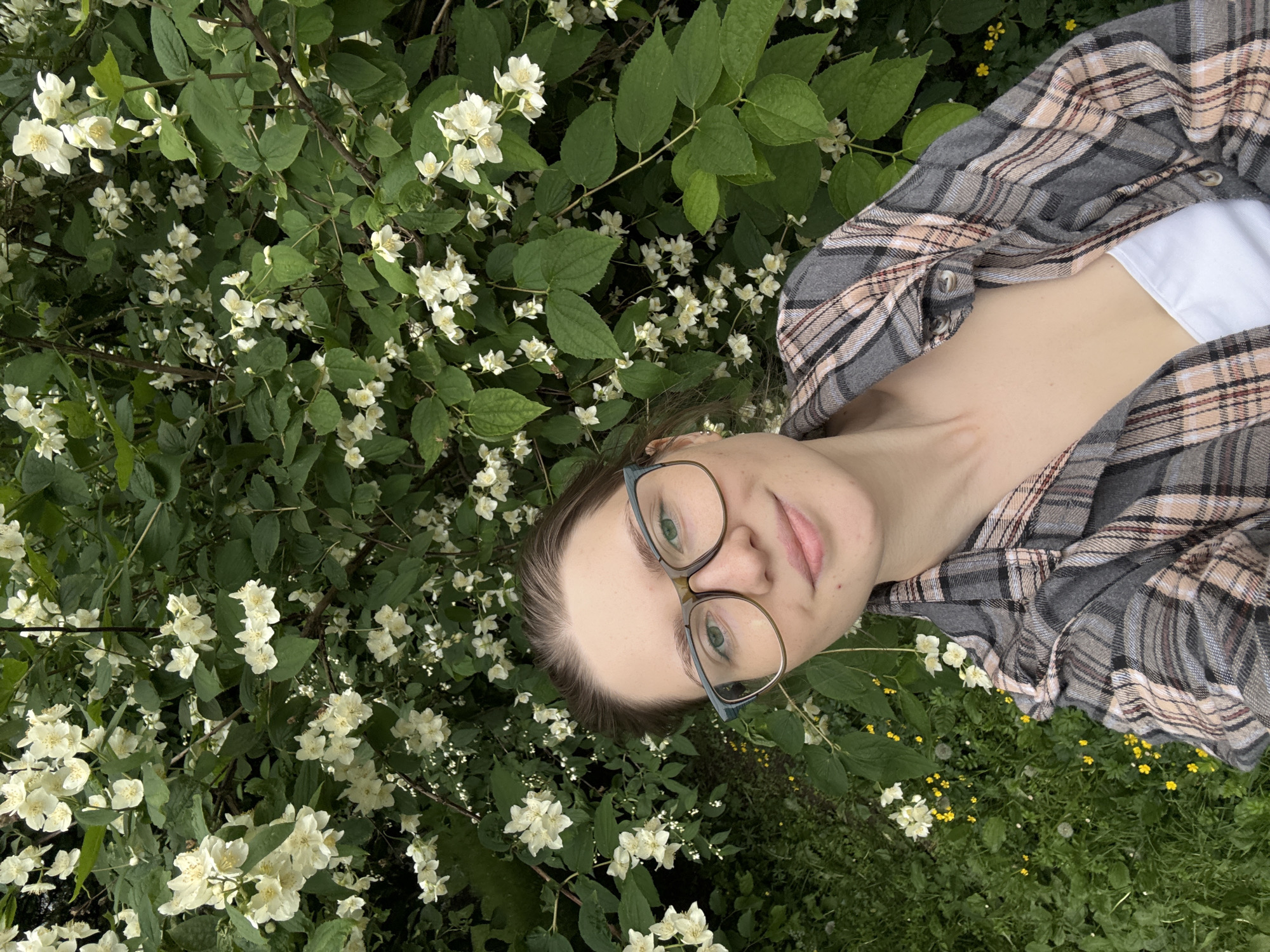 A young woman with brown hair pulled back, wearing glasses with thick frames and a plaid shirt over a white top, is taking a selfie in front of a bush covered in small white flowers and green leaves. The background is a dense arrangement of foliage, with some small yellow flowers and green grass visible at the bottom left. The lighting suggests an outdoor setting, possibly in a park or garden. No specific location in Saint Petersburg is identifiable from the image.