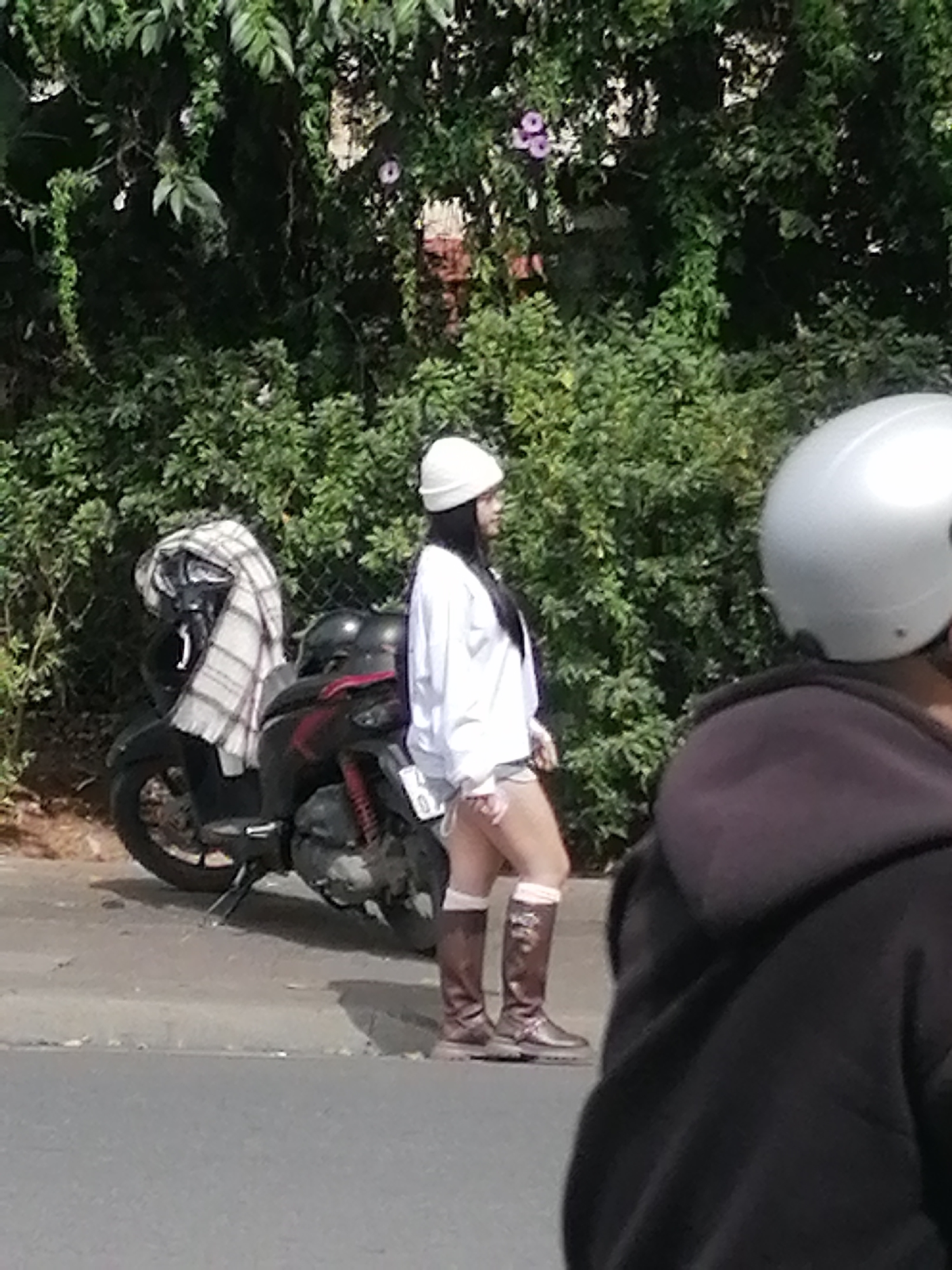 In Da Lat, Vietnam, a woman wearing a white cap and a white shirt stands next to a parked scooter. The scooter has a plaid blanket draped over its handlebars. The woman is wearing brown boots and white socks. In the foreground, another person is visible from behind, wearing a dark hoodie and a silver helmet. The background is filled with lush green foliage, suggesting a location with abundant vegetation, common in Da Lat's hilly terrain. The scene appears to be on a roadside, with a paved surface for the road and a grey concrete curb.