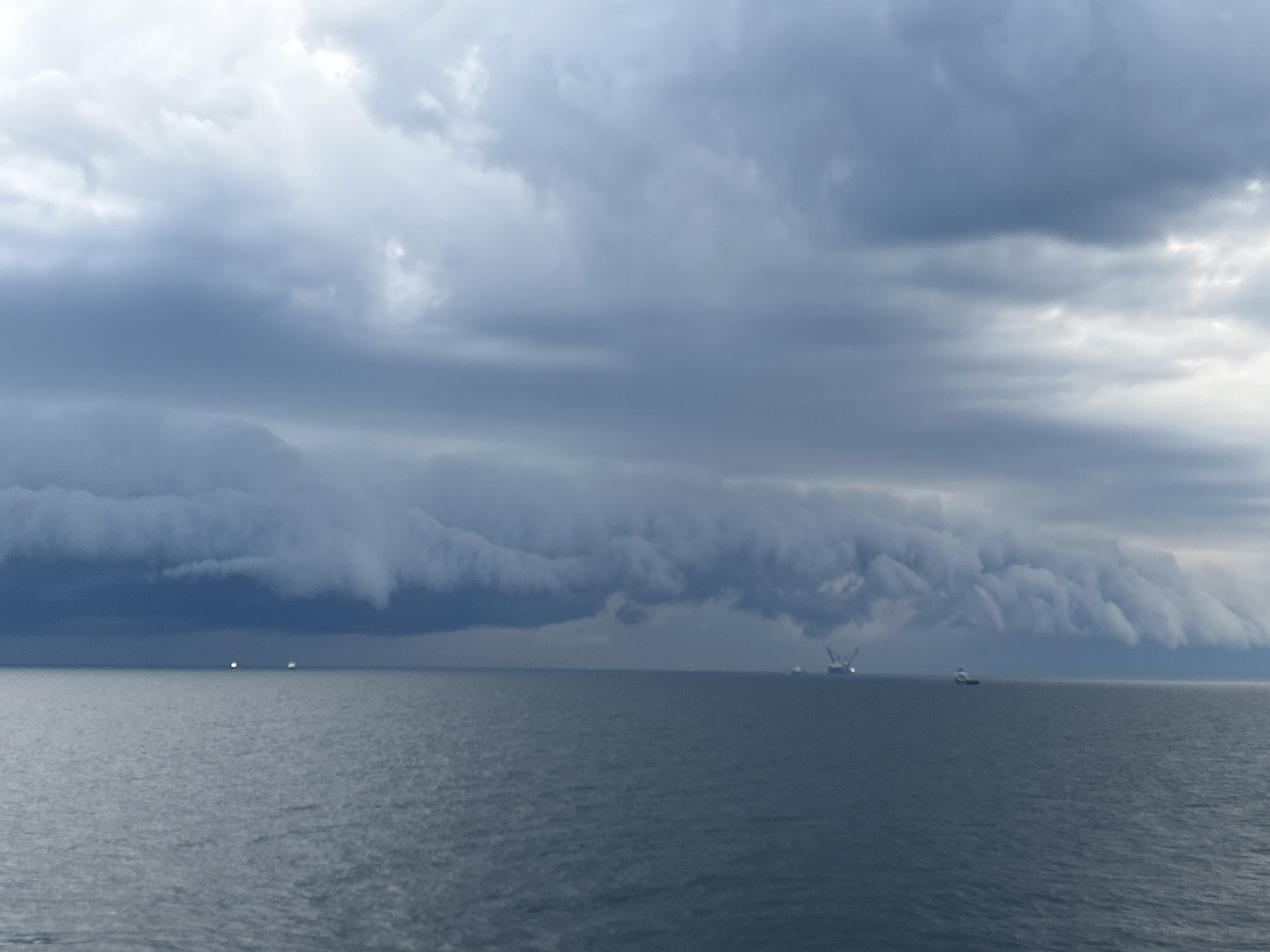 The image captures a vast expanse of dark, calm ocean under a dramatic, overcast sky. The dominant feature is a massive, dark cloud formation stretching horizontally across the horizon, resembling a shelf cloud or a powerful storm front. This cloud band is dark blue-grey and appears turbulent and heavy, with lighter patches of grey and white clouds visible in the upper sky above it.
On the dark water, several objects are discernible along the horizon line. Towards the left, two small, bright white lights are visible, possibly from distant vessels or buoys. In the middle-right, a larger industrial structure, consistent with an offshore oil rig or a large work platform, is present, showing some visible lights and crane-like elements. Further to the right, closer to the foreground, a smaller white boat or vessel is visible on the water.
The scene depicts pre-storm or stormy weather conditions, with the heavy cloud cover creating a somber, low-light atmosphere, suggesting it is either a very dark day or approaching twilight. Despite the menacing sky, the water's surface appears relatively calm. There is no visible text in the image. The overall impression is one of natural grandeur and the imposing power of weather over the open sea, likely in a marine industrial setting.