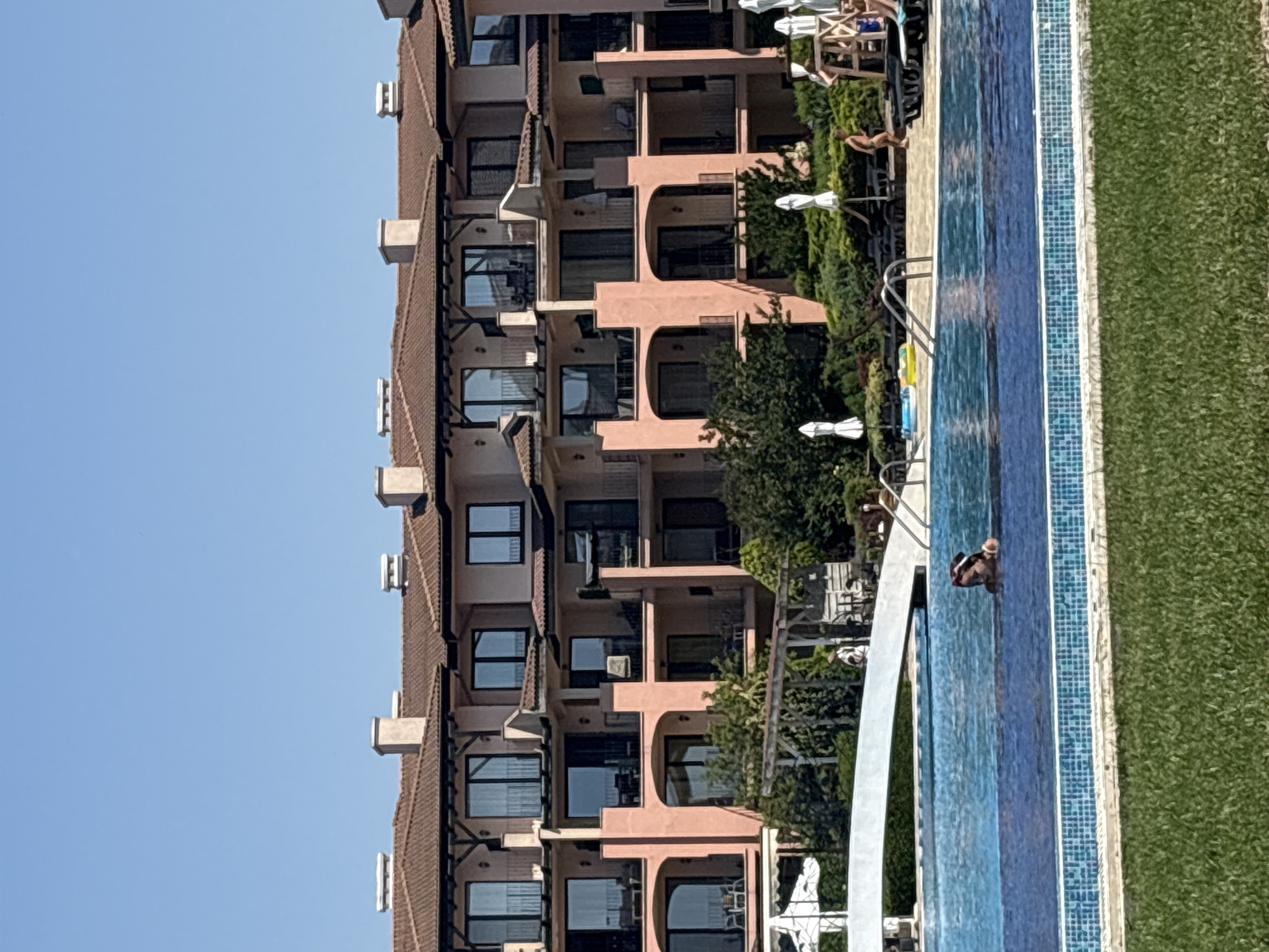 A wide shot captures a swimming pool in the foreground with a woman partially submerged in the water. Beyond the pool, a white arched bridge spans across a section of it. Further in the background, a multi-story pink building with red-tiled roofs and numerous balconies is visible. Several white umbrellas are dotted around the pool area, and lush green bushes and trees surround the building and pool. The sky is clear and blue.