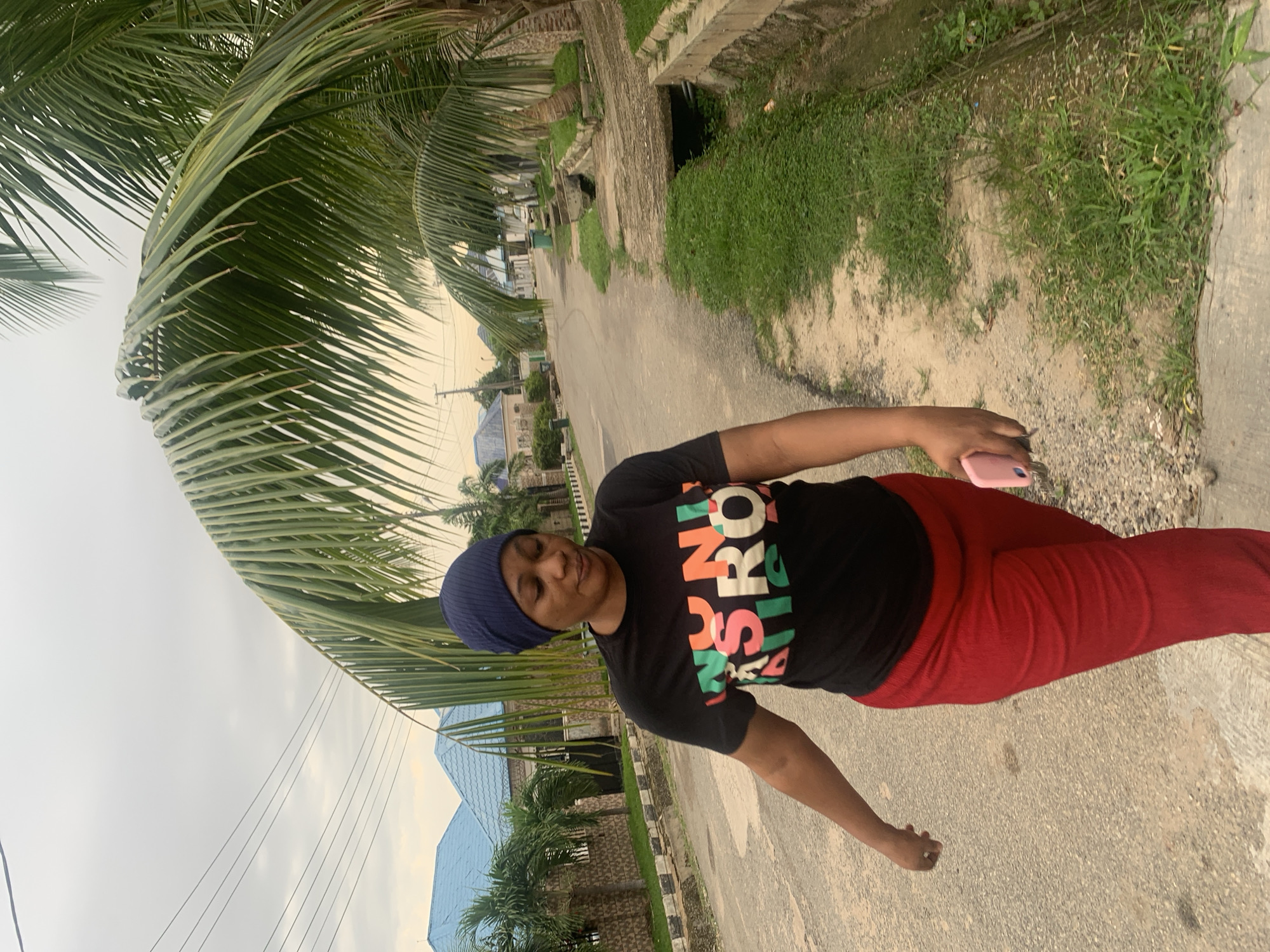 A woman with dark skin wears a dark blue headscarf and a black t-shirt with colorful lettering. She is wearing red pants. The woman walks along a paved road in a suburban area of Abuja, Nigeria. Houses with blue roofs line the street, and palm trees are visible. The sky is overcast.