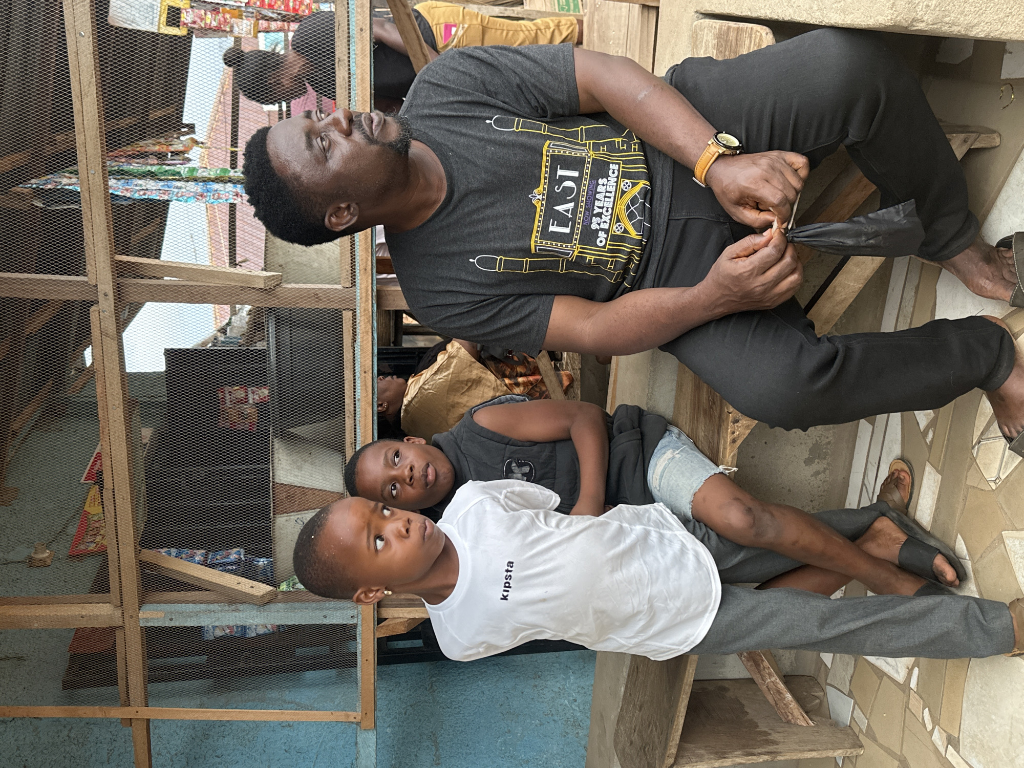 In Narayi, Nigeria, a man and two young boys are seated on a wooden bench in front of a structure with a wire mesh enclosure. The man, on the right, wears a dark t-shirt with a yellow and white graphic and black pants. He has a beard and is looking to his right, holding a small black object in his hands. To his left, two boys are seated. The boy on the far left wears a white t-shirt with the word "kipsta" and gray pants. The boy next to him wears a dark vest over a t-shirt and light-colored shorts. Both boys are looking to their left. Behind them, the wire mesh enclosure reveals shelves with various items, possibly goods for sale. The background shows glimpses of other structures and vegetation, suggesting an outdoor, urban or semi-urban environment. The ground in front of the bench is paved with beige and brown tiles arranged in a mosaic pattern.
