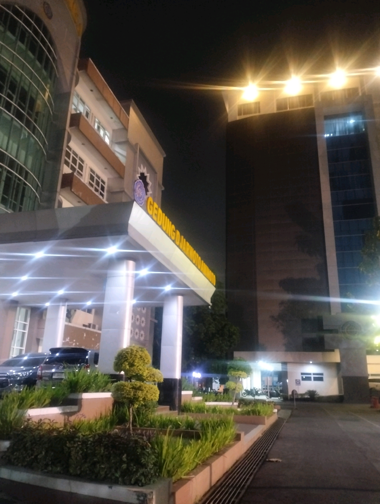 The image depicts the exterior of a modern building at night, likely in Karangsoka, Indonesia. The building features a curved glass facade on the left and a darker, taller structure on the right. In the foreground, a covered entrance area with bright, star-shaped lights is visible. There is landscaping with manicured bushes and trees, and two cars parked in the background. Further to the right, a smaller building with lit windows and several motorcycles parked in front is also present. The overall scene suggests an institutional or commercial building illuminated at night.