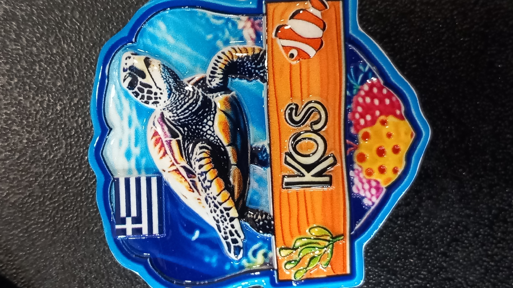 This is a souvenir magnet depicting a sea turtle swimming underwater with a Greek flag in the background. Text on the magnet reads "Kos." The image is set against a textured black background. The magnet has a blue, wavy border. The scene includes coral and a clownfish. The magnet is of the island of Kos, Greece.