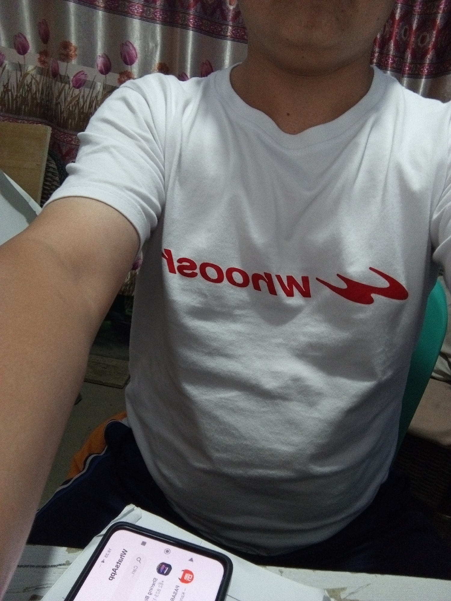 A person is wearing a white t-shirt with a red graphic logo on it. The person is seated in a room with patterned curtains in the background. A smartphone is visible in the foreground. The scene does not provide any specific information or context to determine its location within the city of Cihanjuang, Indonesia.