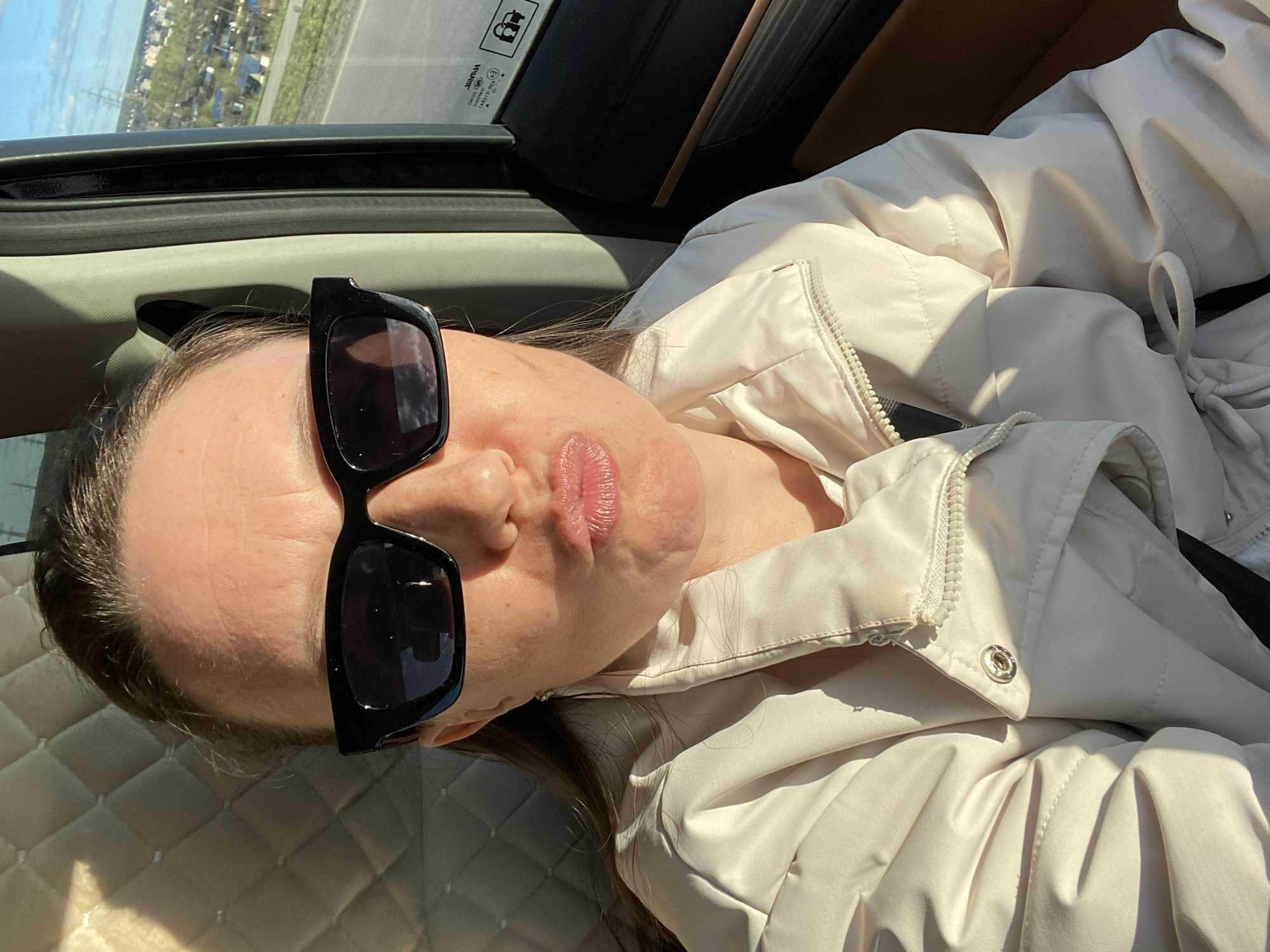 A woman with sunglasses is lying in a car. The location is Chaikovsky, Russia, as indicated by the license plate on the window. She is wearing a white jacket. The car has a sunroof. The seat is light colored and quilted.