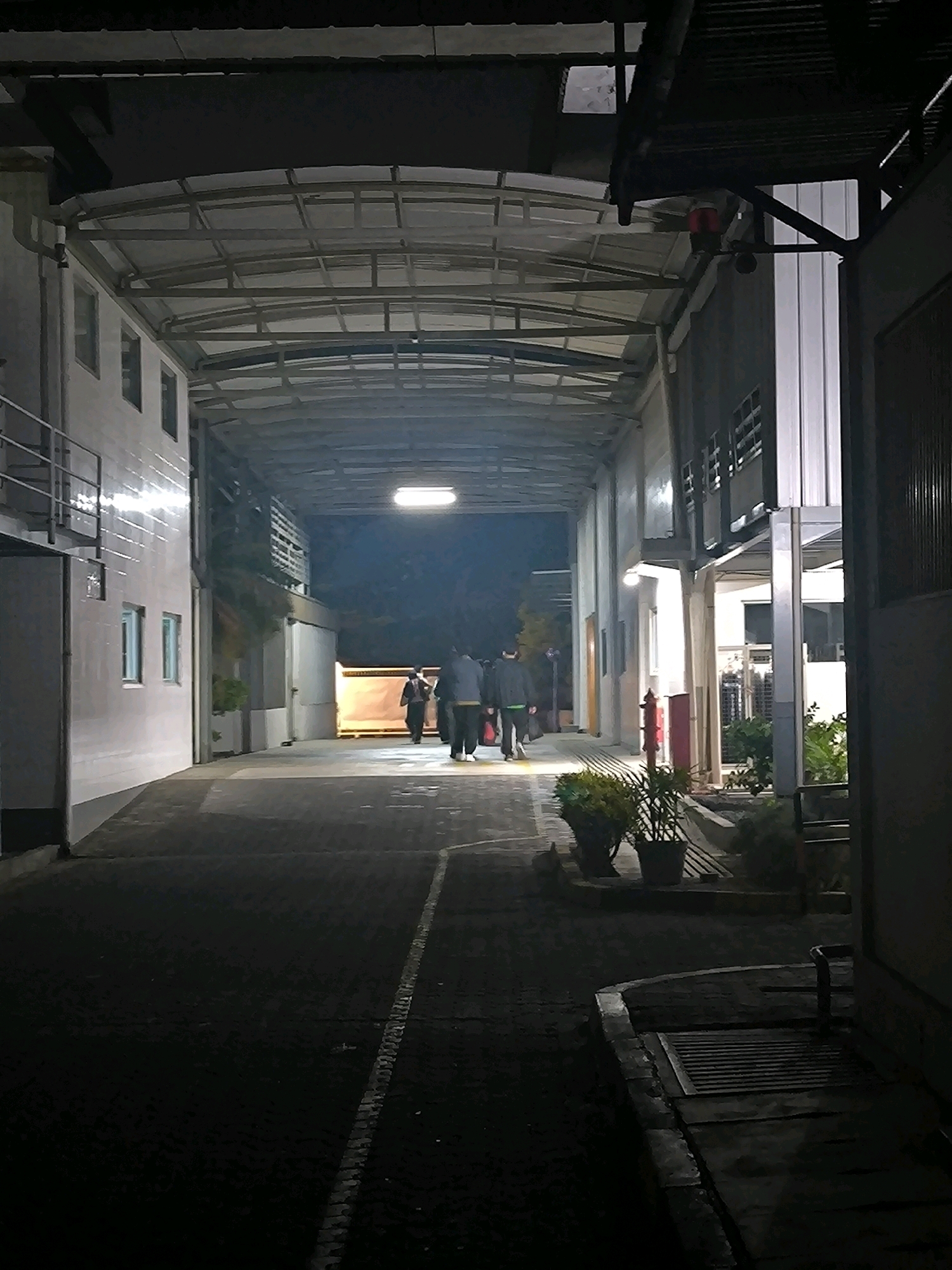 The image depicts a covered walkway at night, likely in an industrial or commercial area. The walkway is illuminated by overhead fluorescent lights and appears to be paved with interlocking concrete blocks. On the left, a white building with a metal railing runs along the side. On the right, another building is visible, featuring a corrugated metal facade and a section with vertical white panels. Two people are walking away from the camera down the walkway. A third person, wearing a yellow jacket, is further ahead and is partially obscured. In the foreground on the right, there are potted plants and a red fire hydrant. The scene is dark, suggesting it is late at night. The architecture and surrounding environment do not provide specific clues to pinpoint the location within Bandung City, Indonesia.