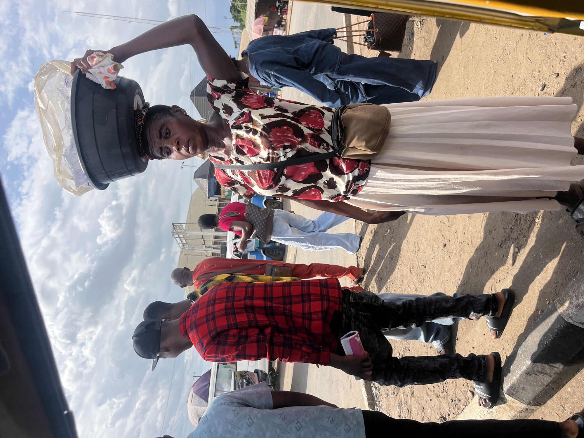 A medium, outdoor shot depicts a group of people in Unknown. The image is somewhat tilted. An African woman balances a large black tub on her head. The tub is covered with a white plastic bag. She wears a dark brown and white patterned shirt, a light-colored skirt, and a brown purse slung across her body. A line of people stand in front of her, including a man in a red and black checkered shirt, dark pants, a black cap, and black sandals. He holds a pink object in his left hand. Others are obscured by those in the foreground, but they appear to be standing on the side of a street. The sky is overcast.