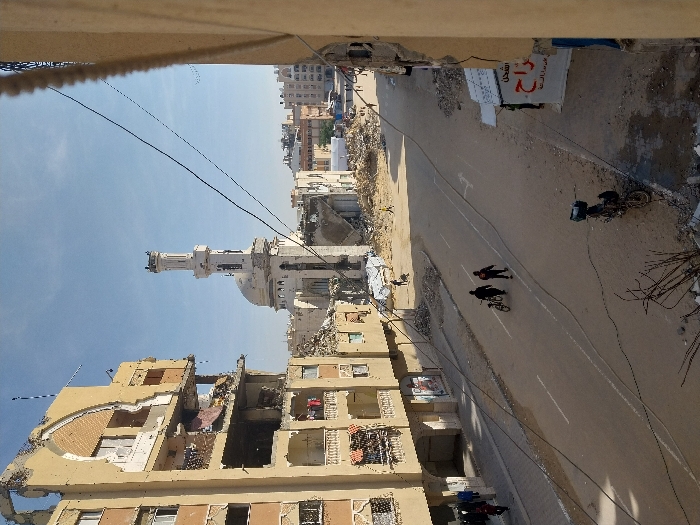 The image depicts a devastated urban street scene in Khan Yunis, Palestinian Territory, under a clear blue sky. In the foreground, spanning the bottom of the frame, are debris, bare branches, and multiple dark wires crossing diagonally. A motorcycle is parked on the dirt shoulder to the right. The midground features a two-lane paved road, partially covered in dust and debris, with faded white lane markings. Two individuals dressed in dark clothing walk away from the viewer along the left side of the road, one pushing a bicycle.
To the left of the road stands a heavily damaged multi-story apartment building, light beige in color, with significant structural collapse revealing interior rooms. Laundry hangs from some of its remaining balconies. To the right of the road are extensive piles of rubble and debris, with a white awning featuring Arabic script visible over a small structure.
The background is dominated by a mosque with a light-colored dome and a tall, slender minaret, both exhibiting visible structural damage, particularly at the base of the dome and surrounding structures. Beyond the mosque, more damaged buildings and rubble piles extend into the distance. Several small figures are discernible walking through the rubble further down the street. The overall environment indicates widespread destruction and civilian presence amidst the damage.
