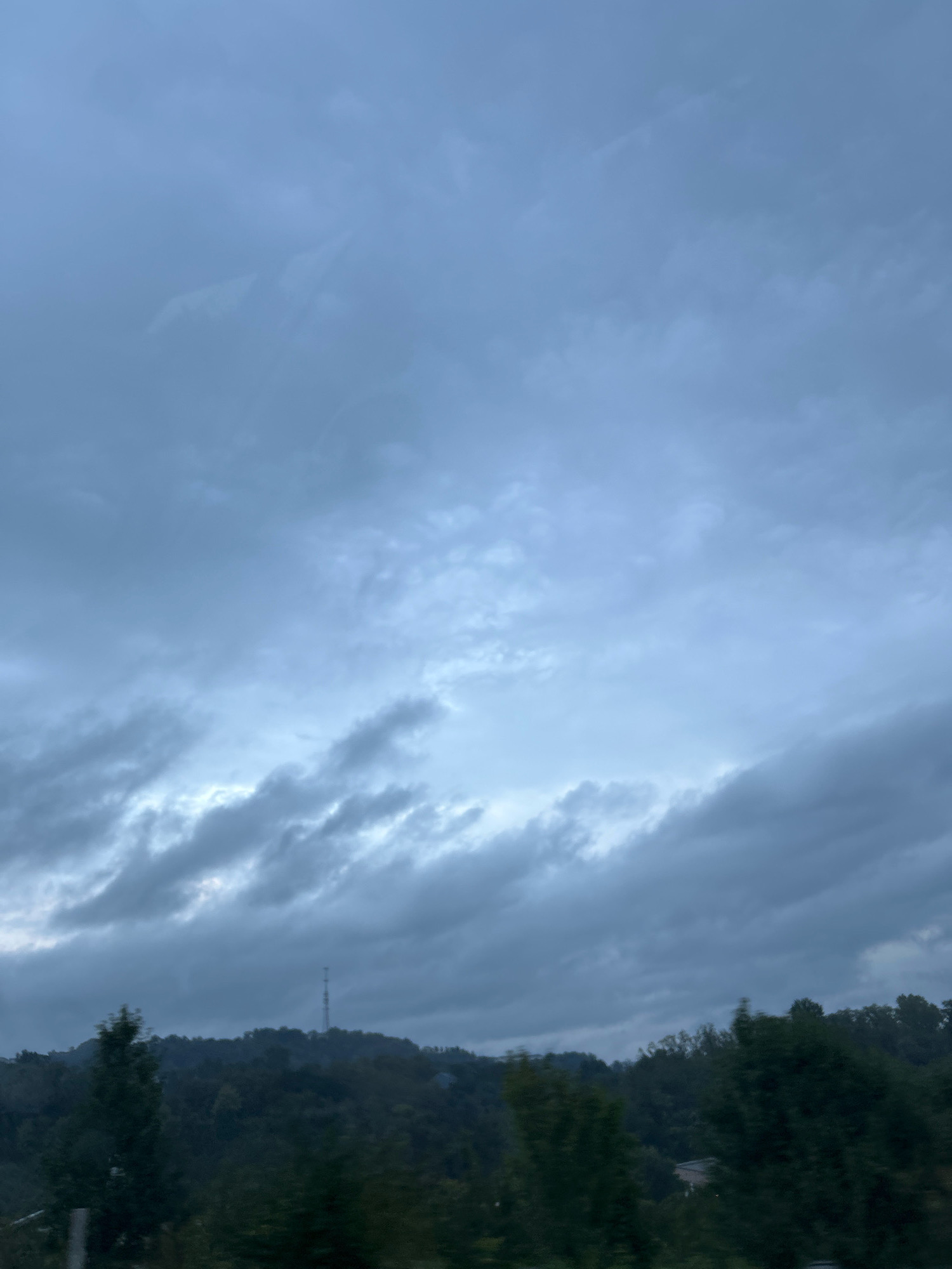 A telecommunications tower stands on a tree-covered hill under a cloudy sky. The sky is filled with various cloud formations, ranging from wispy to thick and dark. Below the sky, a hilly landscape is visible, densely covered with trees. The tower is positioned near the crest of the hill, and it appears to be a tall, slender structure. The overall scene suggests an overcast or impending stormy weather condition. The location is identified as Collier Township, United States of America.