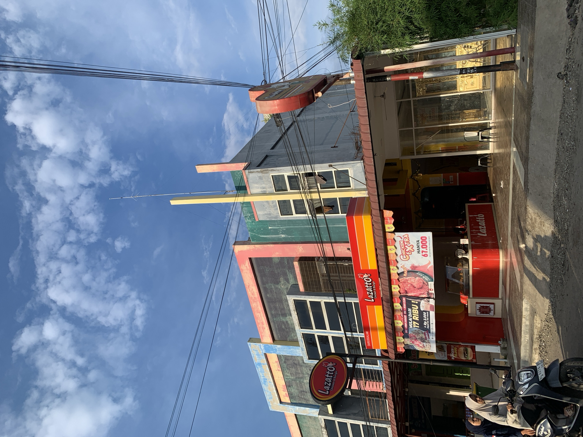 The image shows a street view in Lhokseumawe, Indonesia, featuring a multi-story building with a colorful facade, housing a "Lazatto Chicken & Burger" establishment. The building's upper stories are painted in a mix of blue, pink, and green, with large, dark-tinted windows. Below, the Lazatto restaurant has a prominent red and yellow awning with signage. Inside the restaurant, a display counter showcases fried chicken. Two individuals are riding a motorcycle on the street in front of the building, heading towards the viewer. Overhead, a complex network of power lines is visible against a bright blue sky with scattered clouds. The ground appears to be a paved street with some gravel.