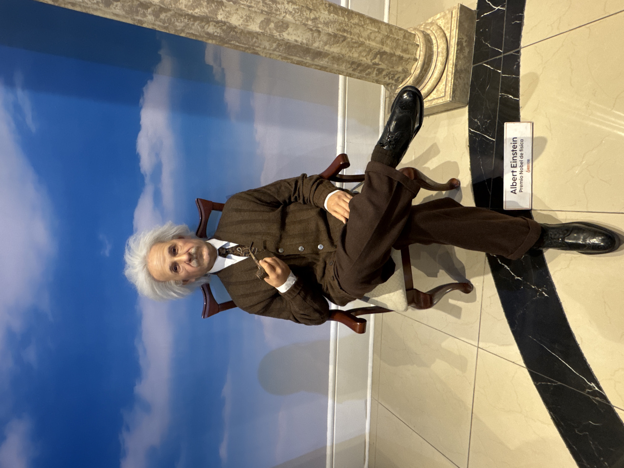 The image shows a wax figure of Albert Einstein sitting in a chair with one leg propped up. He is wearing a brown striped suit and holding a pipe. A sign next to the figure identifies him as Albert Einstein, Premio Nobel de Física. Behind the figure is a painted backdrop of a blue sky with clouds. The figure is located at the Museo de Cera (Wax Museum) in Mexico City, Mexico. The floor is tiled in alternating light beige and black marble-patterned tiles. The picture shows a column structure and part of the white wall.