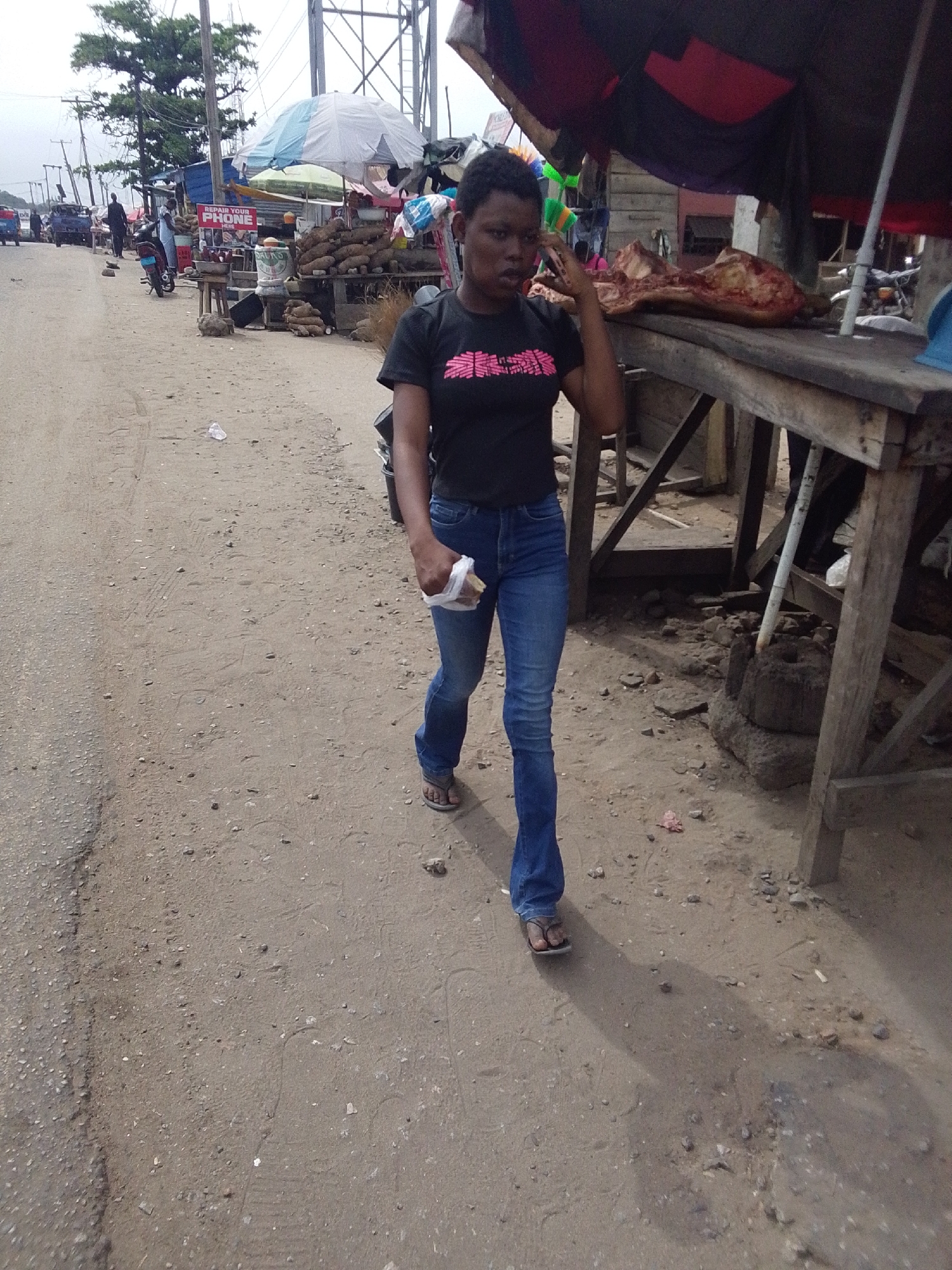 A woman with short black hair, wearing a black t-shirt with pink writing and blue jeans, walks on a dirt road. She holds a small white bag in her left hand and has her right hand raised to her ear as if on a phone call. To her right is a wooden table with a large piece of meat on it. Behind the table is a makeshift awning. Further down the road are vendors selling yams and other produce under an umbrella. A sign that says "REPAIR YOUR PHONE HERE" is also visible. There are people and vehicles in the distance. The ground is dirt and gravel. The scene appears to be in an informal market area in Lokoja, Nigeria.