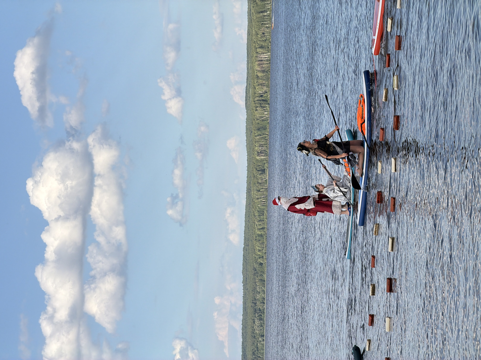 The image depicts a body of water, likely a lake or river, with a dense forest in the background. The sky is blue with scattered white clouds. In the foreground, several people are on paddleboards. One person is dressed as Santa Claus, standing on a paddleboard. Another person, dressed in a black outfit with a headpiece, is kneeling on a paddleboard and holding a paddle. A third person in a light-colored garment is seated on a paddleboard between the Santa figure and the person in black. There are several floating markers in the water. The scene appears to be in Chaikovsky, Russia, given the presence of a boat on the horizon that resembles a boat often seen in the Kama River, which flows through Chaikovsky.