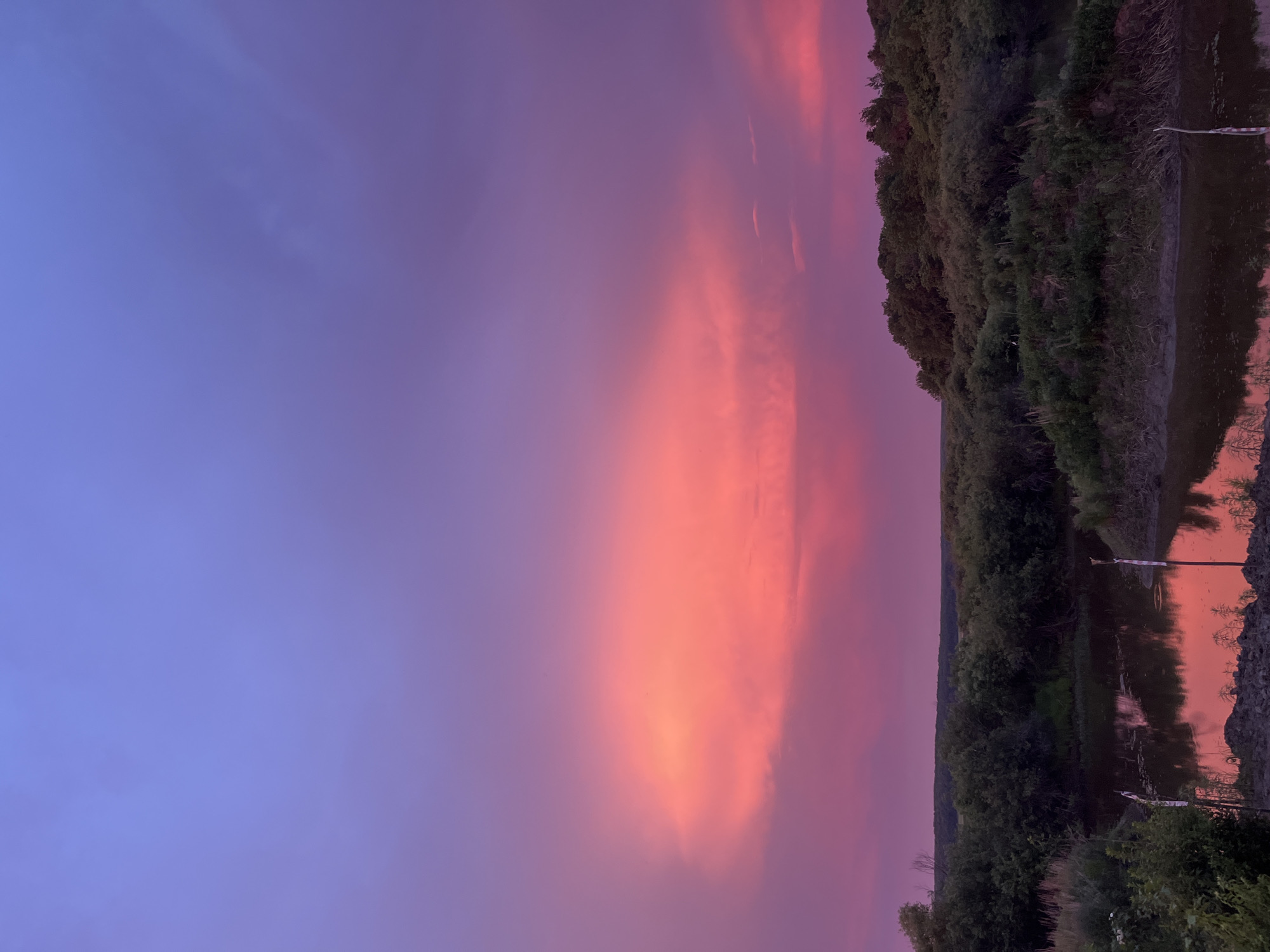 The image depicts a serene natural landscape at dusk, with a vibrant sky painted in hues of purple, pink, and orange. A body of water, possibly a river or lake, occupies the lower portion of the frame, reflecting the colorful sky. Lush green vegetation, including trees and bushes, lines the banks of the water and stretches towards the horizon. The sky is dominated by a large, illuminated cloud formation that glows with intense pink and orange light, contrasting with the deeper purple and blue tones of the upper sky. The overall scene suggests a tranquil and picturesque natural setting, likely in a rural or wilderness area. No people or specific urban features are visible in the image.