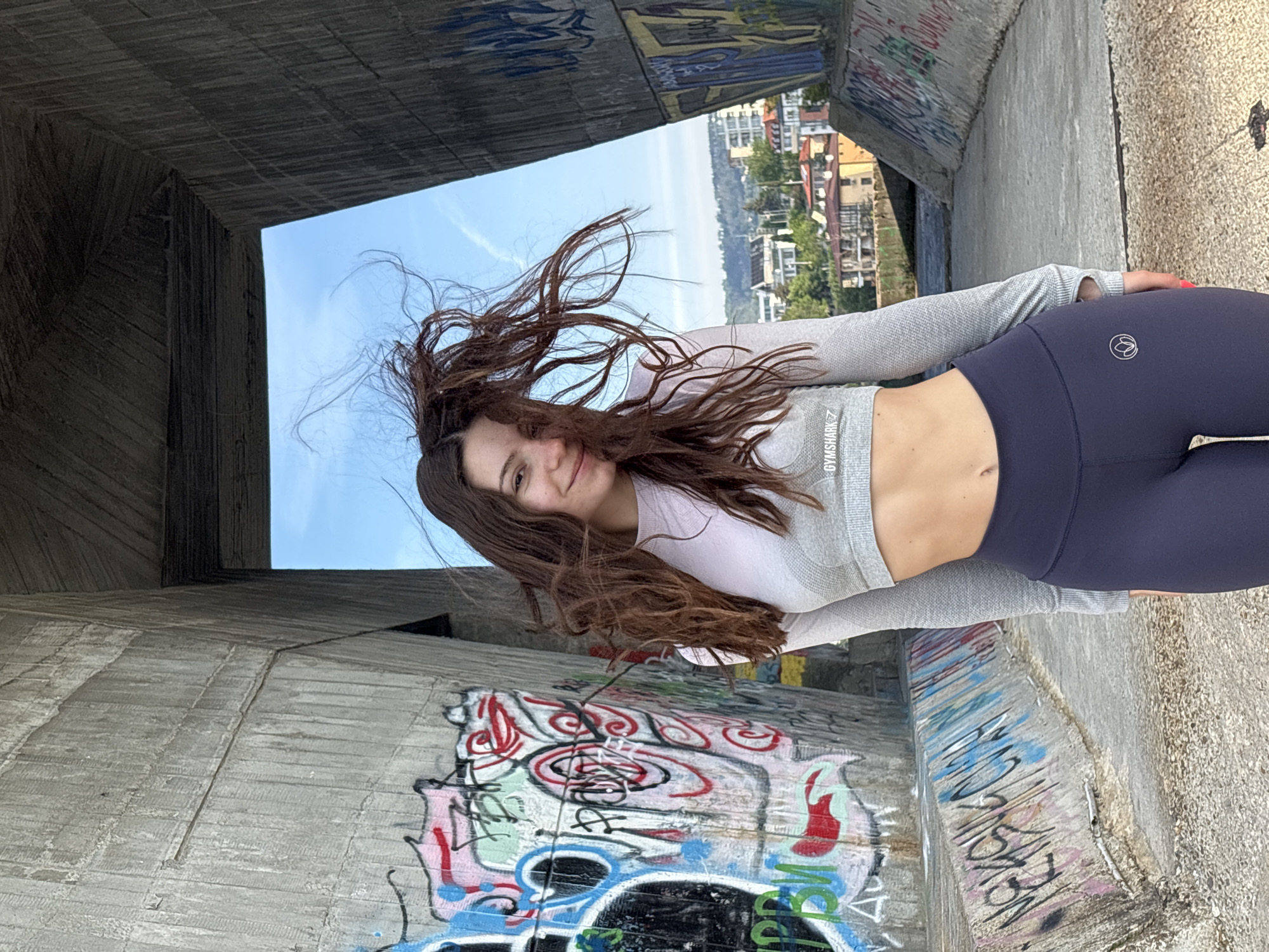 A young woman with brown hair, wearing a grey long-sleeved crop top and dark blue leggings, stands in a concrete tunnel with graffiti-covered walls. The opening of the tunnel provides a view of buildings and a blue sky, suggesting an urban setting. The woman is smiling, and her hair is blowing in the wind. The location is likely in Varna, Bulgaria, indicated by the urban landscape visible through the tunnel opening.
