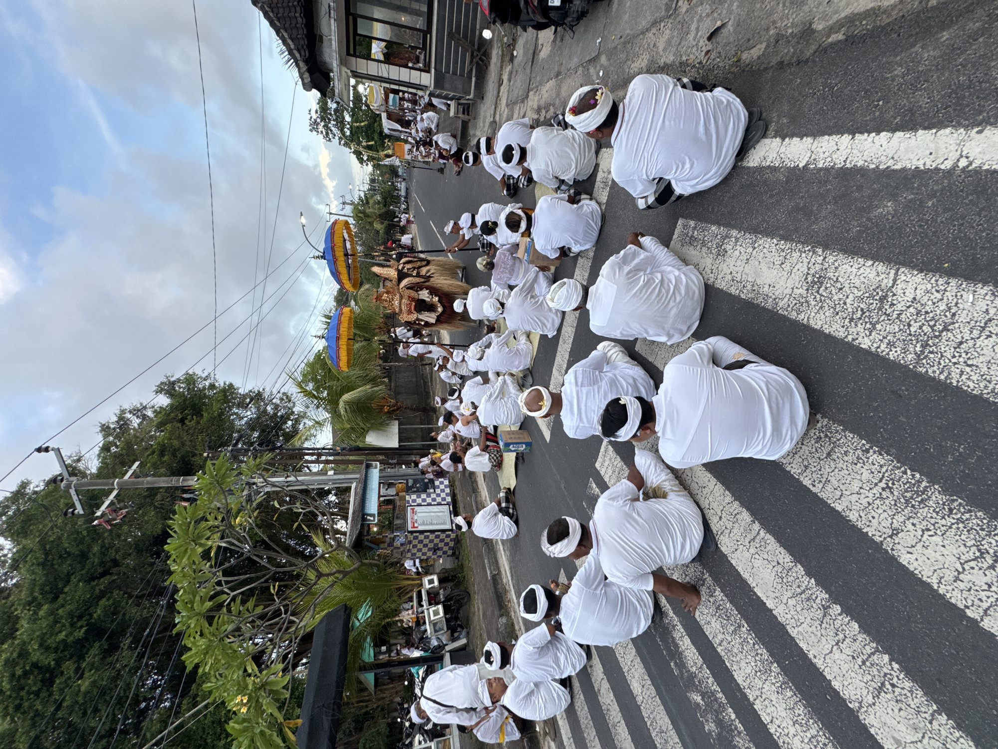 A street scene in Kuta, Indonesia shows a large group of people kneeling on a crosswalk. All are wearing white garments, many also wearing white head wraps. An elaborately decorated, golden-colored palanquin is being carried along the street, preceded by two yellow and blue umbrellas on poles. Trees and buildings line the street, and overhead electrical wires are visible against a cloudy sky.