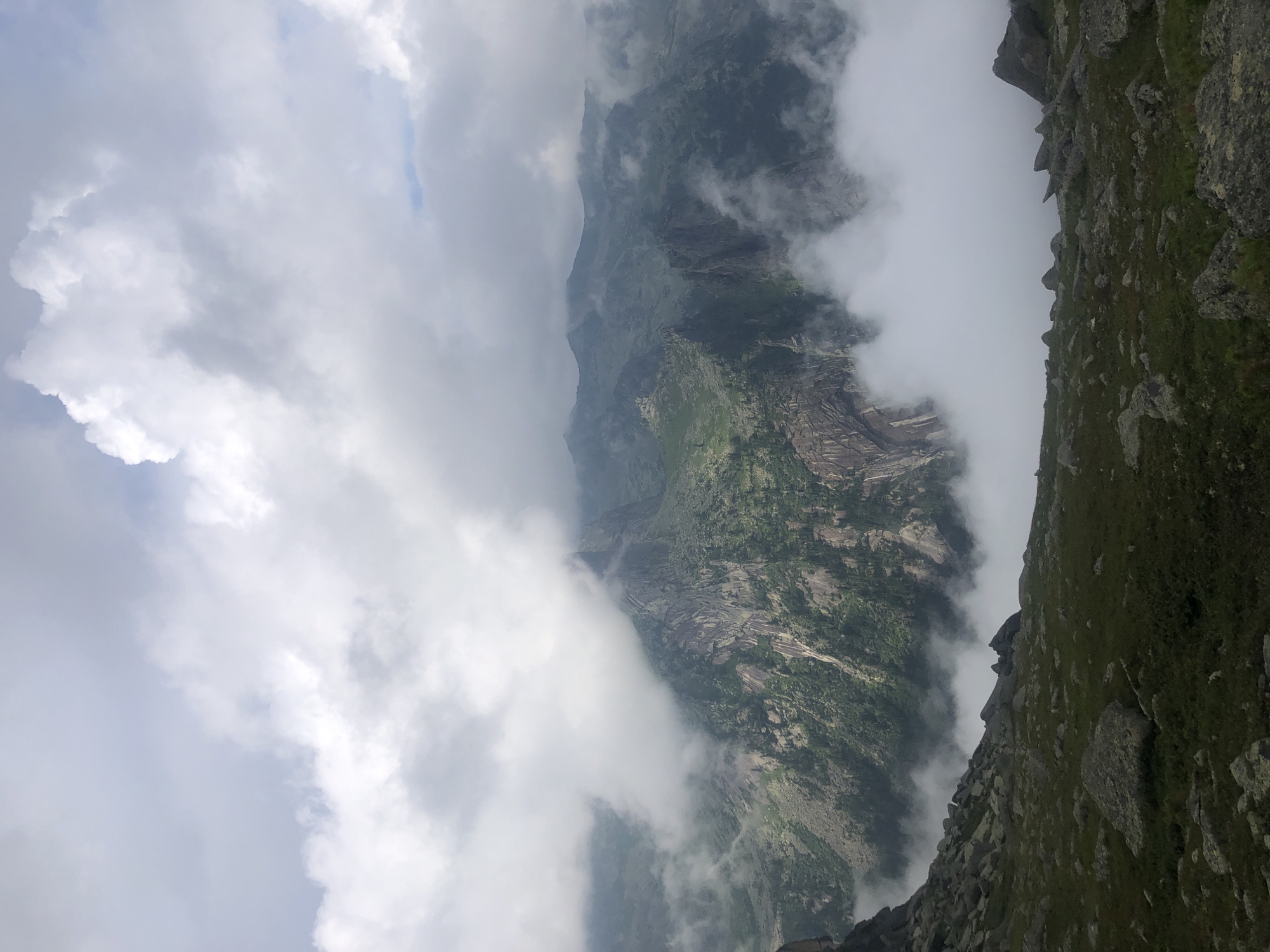The image shows a mountainous landscape with clouds partially obscuring the view. In the foreground, there is a rocky, grassy slope. The middle ground features steep, rocky mountain faces with patches of green vegetation, likely trees and shrubs, clinging to the slopes. Further in the background, a range of mountains recedes into the distance, with some peaks also covered by clouds. The sky is filled with dramatic, textured clouds, suggesting a dynamic weather system. The overall impression is one of rugged natural beauty and a sense of elevation. The location is identified as Ergaki Nature Park, Krasnoyarsk Krai, Russia.