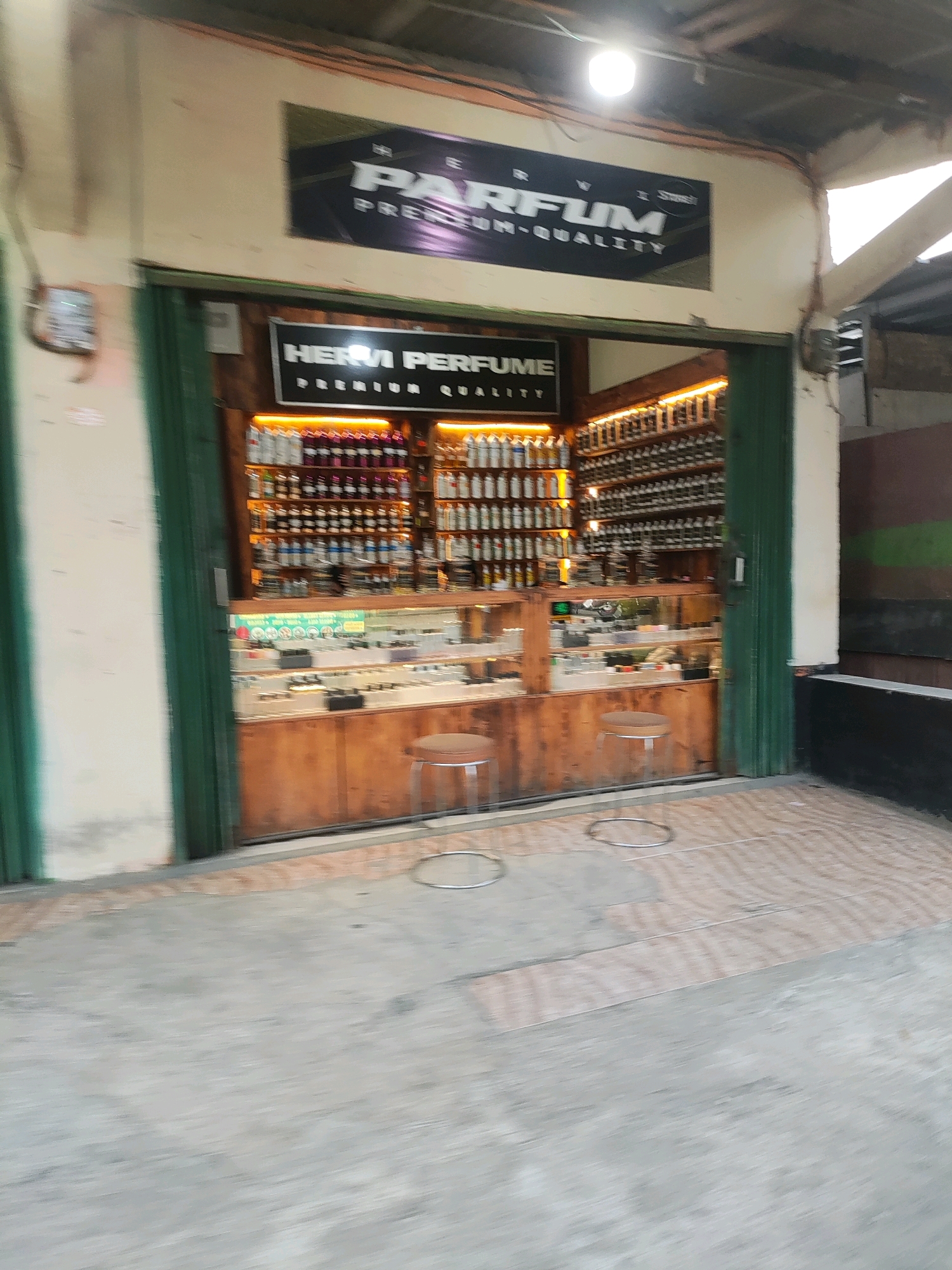 The image shows the exterior of a perfume shop located in Wanajaya, Indonesia. The shop has a large open entrance with green-painted folding doors. Above the entrance, there are two signs. The top sign is black with white lettering and appears to read "PARFUM PREMIUM-QUALITY". Below this, a smaller sign inside the shop reads "HERVI PERFUME PREMIUM QUALITY".
The interior of the shop is well-lit and features rows and rows of small bottles, presumably containing perfumes, displayed on shelves. The shelves are made of wood and are illuminated from above. There is also a glass display case in front of the shelves, showing more small items. Two round stools with metal legs and wooden seats are placed outside the shop entrance on the patterned concrete floor. The floor is a mix of concrete and what appears to be wooden planks. The overall impression is a small retail establishment specializing in perfumes.