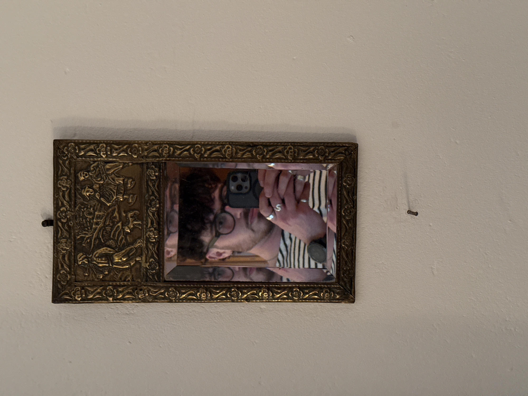 A tarnished gold-colored rectangular mirror with ornate detailing is affixed to a pale, off-white wall. A reflection of a person wearing glasses and a striped shirt can be seen in the mirror, and they are holding a smartphone pointed towards the mirror, presumably taking a picture. A small metal fastener is visible to the right of the mirror. The location is Unknown, Unknown.