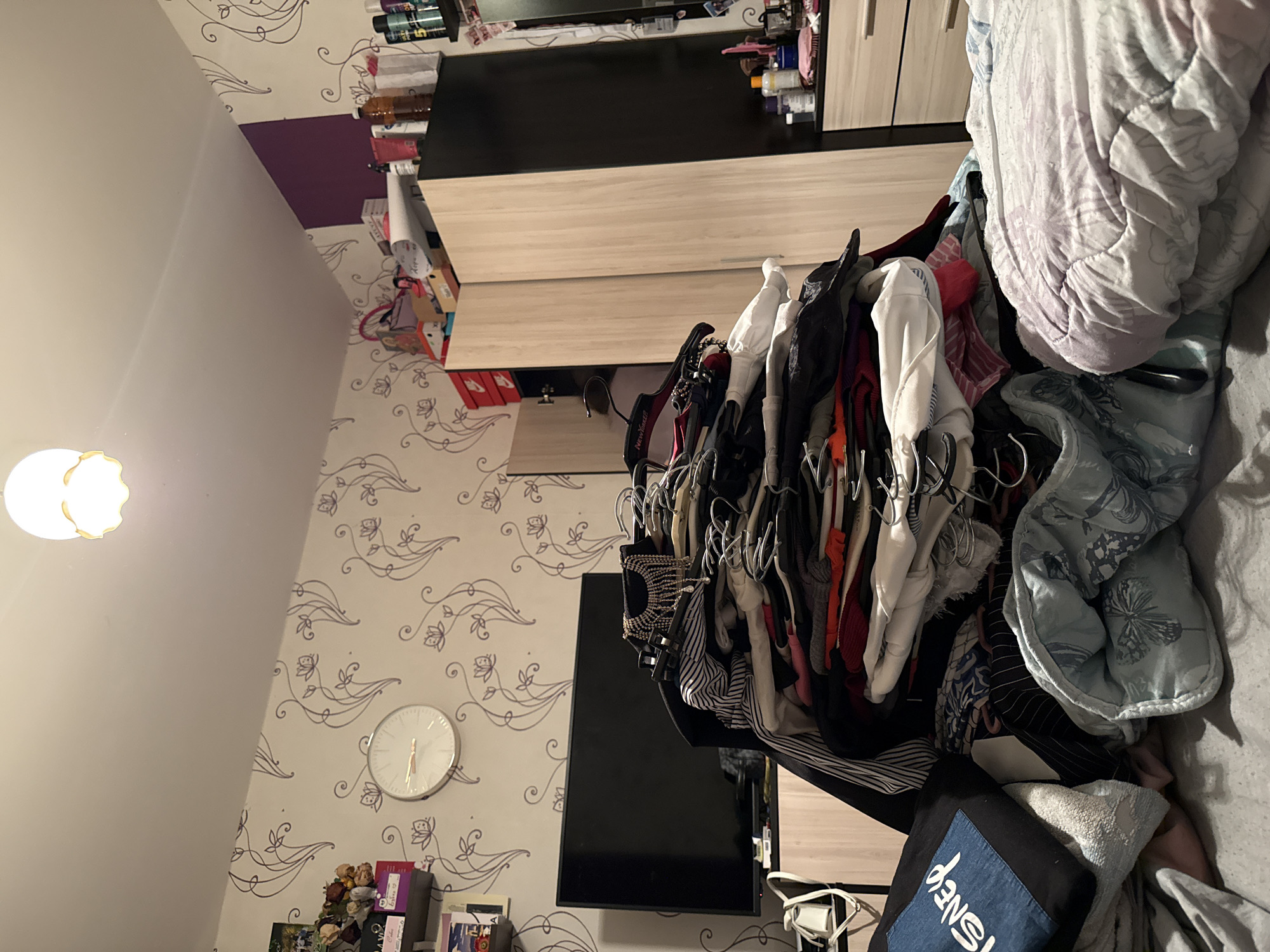 The image depicts an indoor scene, likely a bedroom, characterized by clutter. A large, disorganized pile of clothing is the central focus, occupying the foreground and midground. The clothing appears to be a mix of garments and hangers, with various colors and textures visible. To the left of the clothing pile, a black television screen is mounted on a stand. Behind the television, a wall is adorned with patterned wallpaper featuring swirling floral designs. A circular clock with a white face and black hands hangs on the wall above the television.
To the right of the clothing pile, two large wardrobes are visible. The wardrobe on the left has light-colored doors, while the wardrobe on the right is predominantly black with a lighter section. The top of the wardrobes is cluttered with various items, including boxes and bottles. A purple section of the wall is visible to the right of the wardrobe.
In the foreground, a bed or sofa is partially visible, covered with bedding. A black tote bag with the word "DISNEY" printed in white on a blue patch is placed on top of some folded items.
The lighting in the room appears to be artificial, with a ceiling light fixture visible at the top of the image. The overall impression is one of disarray, with emphasis on the piled clothing. The image does not provide any specific visual cues to indicate its location within Stara Zagora, Bulgaria.