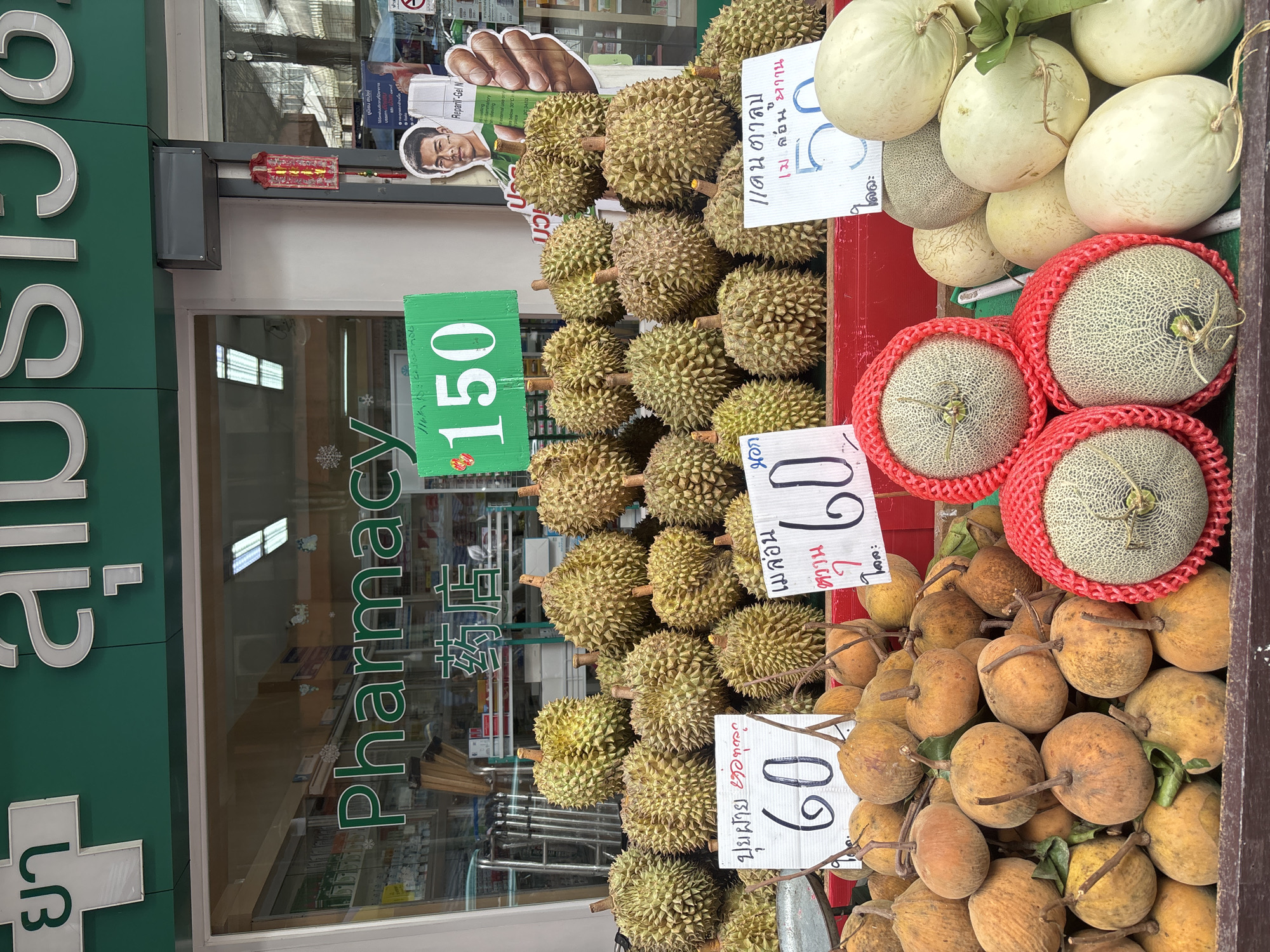 The image depicts a fruit stand in front of a "Pharmacy" store in Chiang Mai City Municipality, Thailand. The pharmacy sign is visible in green and white lettering with Chinese characters below it. The fruit stand displays a variety of fruits, including durians, santols, and cantaloupes. Signs with prices are placed among the fruits. One sign shows "150" in green, and other signs display "60" and "50" with Thai writing. Several durian fruits are piled up, some with price tags indicating "60". Below the durians, a pile of santol fruits is visible. To the right, cantaloupes are displayed, some wrapped in red netting. A man is pictured on an advertisement poster behind the fruits, holding a tube of cream. The overall scene suggests a bustling market environment on a street.