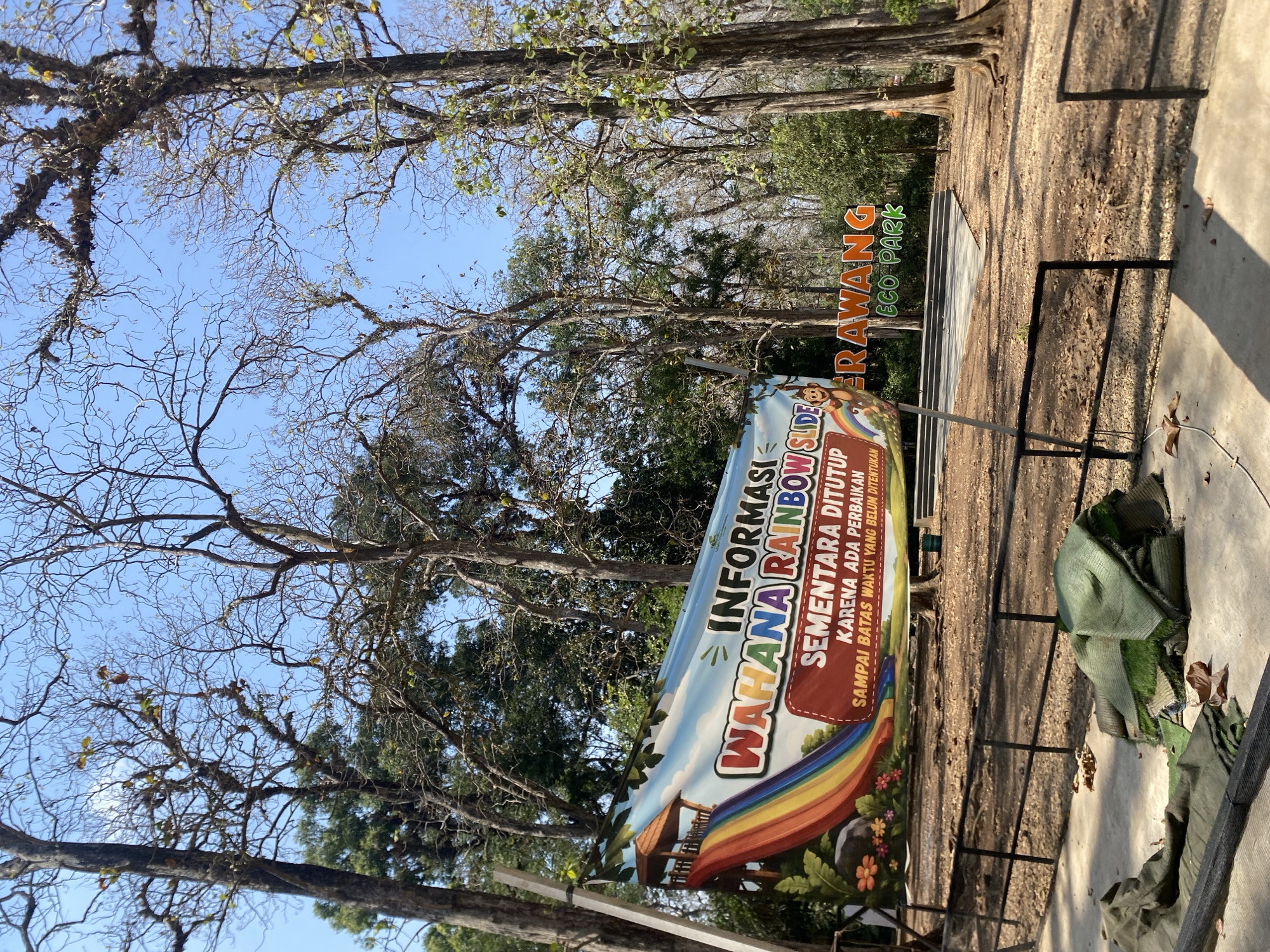 A weathered banner, depicting a rainbow slide and text in Indonesian that translates to "Information: Rainbow Slide Attraction, Currently Closed Due to Maintenance Until Further Notice," is suspended between two poles in front of a clearing. The banner is slightly torn and shows signs of age. Behind the banner, a brightly colored sign reads "KERAWANG ECO PARK." The scene is set within a wooded area with tall, bare trees and a bright blue sky overhead. The ground in the foreground is covered with dirt, fallen leaves, and some rolled-up green material, possibly artificial turf or mats. A black metal fence runs across the midground, separating the viewer from the clearing where the sign is located.