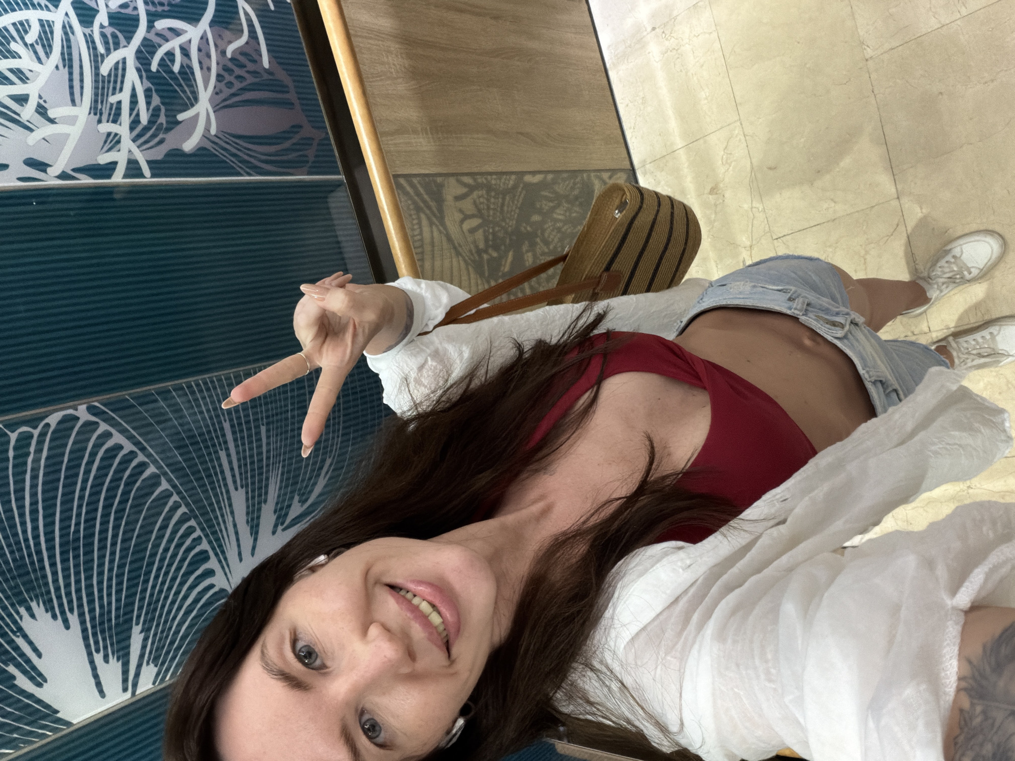 A woman is taking a selfie in what appears to be an indoor setting with patterned walls and a tiled floor. She has long brown hair, blue eyes, and is smiling. She is wearing a red bikini top, denim shorts, a white open shirt, and white sneakers. She is holding her right hand up in a peace sign. A woven straw bag with dark stripes is hanging from her shoulder. The background features teal-colored wall tiles with white leaf-like patterns and wood paneling. There is a wooden handrail visible. The floor is made of light-colored marble tiles. There is no indication of the specific location within Dong Ninh Hoa Ward, Vietnam.