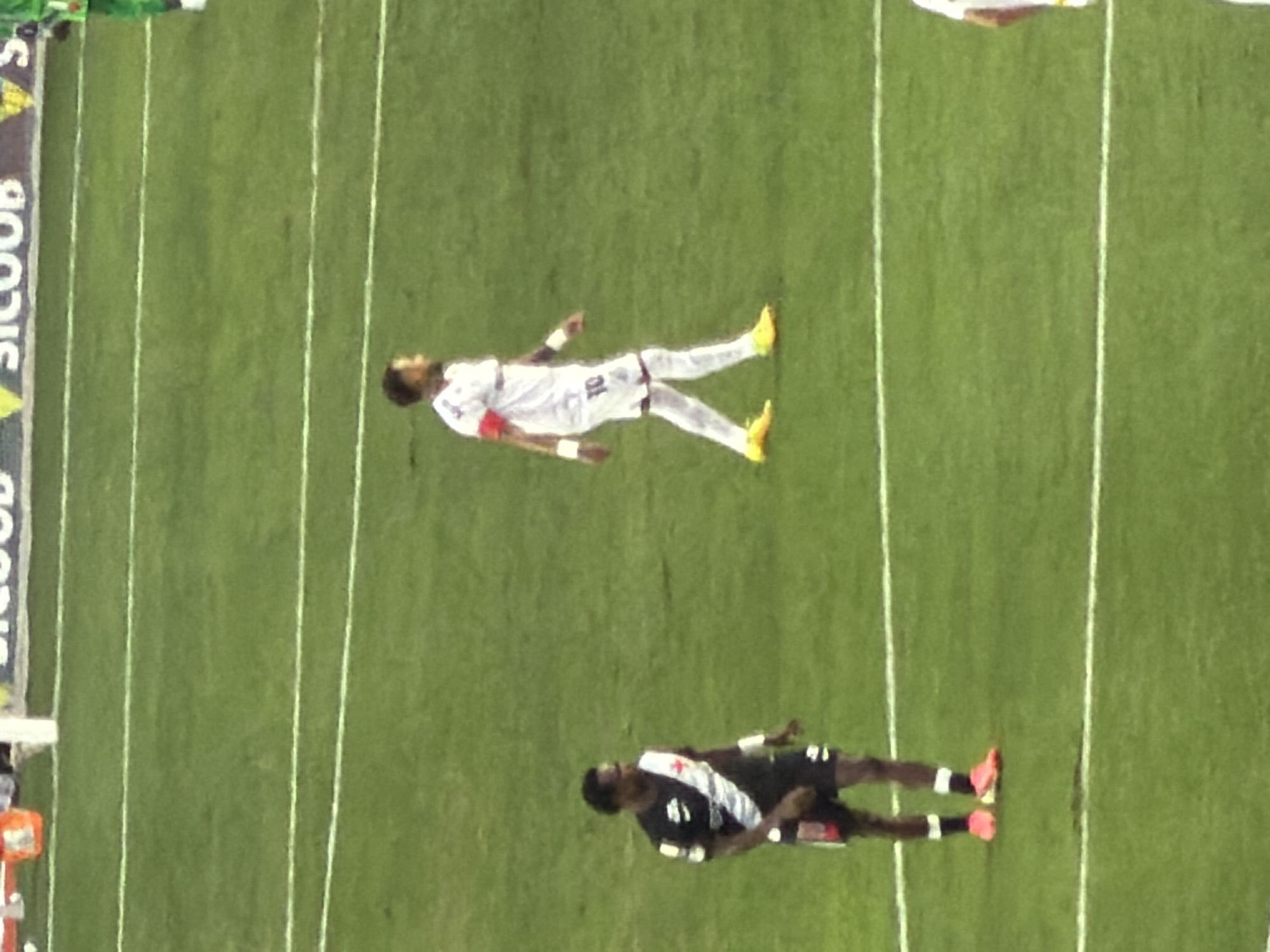 This image depicts a soccer match on a green grass field with white boundary lines. In the foreground, a player wearing a black and white jersey and black shorts stands facing left. To his right, further into the field, another player in a white jersey with the number 10 and white shorts is walking towards the right. The background shows a blurred image of a stadium with advertising boards. The scene is consistent with a professional soccer game played in a stadium, which could be located in Rio de Janeiro, Brazil, given the commonality of such venues in the city. The lighting suggests it is daytime or evening under stadium lights.