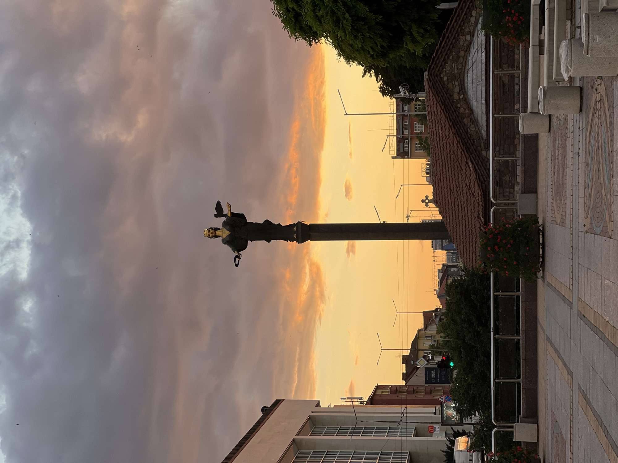 The image depicts the statue of Saint Sophia in Sofia, Bulgaria, during sunset. The statue, made of bronze, stands on a tall column. Saint Sophia is depicted as a woman wearing a crown and holding a laurel wreath in her left hand and a small raven in her right hand. The sky is filled with dramatic clouds, transitioning from grey at the top to vibrant orange and yellow hues near the horizon, indicating the setting sun.
In the foreground, there is a paved plaza with geometric patterns. To the left of the statue is a modern, white building with large, rectangular windows. To the right of the statue, the roof of a small, old building with a red-tiled roof and stone walls is visible. Trees and streetlights are scattered throughout the urban landscape. The scene appears to be a public square or intersection in the city. There are no people visible in the frame, but there are a few small birds flying in the sky. The overall atmosphere is serene and picturesque, enhanced by the warm colors of the sunset.
