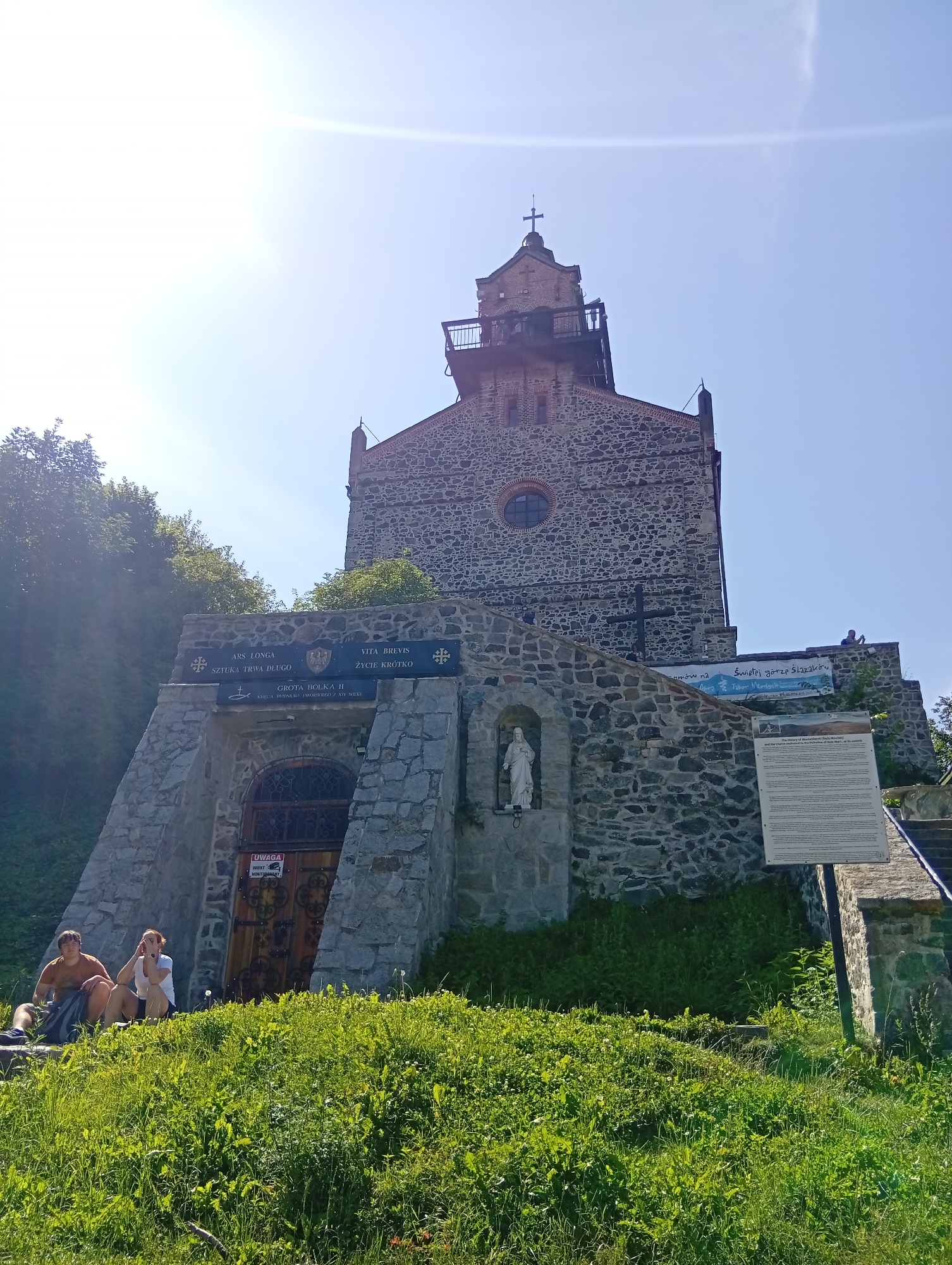 The image shows a stone church atop a hill in Sobótka, Poland. The church has a tower with a cross on top and a balcony. The facade of the church is made of stone, with a circular window and a large wooden door. There are two people sitting on the grass in front of the church, and a statue of a woman in a niche on the side of the building. A sign in Polish and English provides information about the history of the church and the mountain. The sky is clear and blue, and the sun is shining. The church appears to be a popular tourist destination.