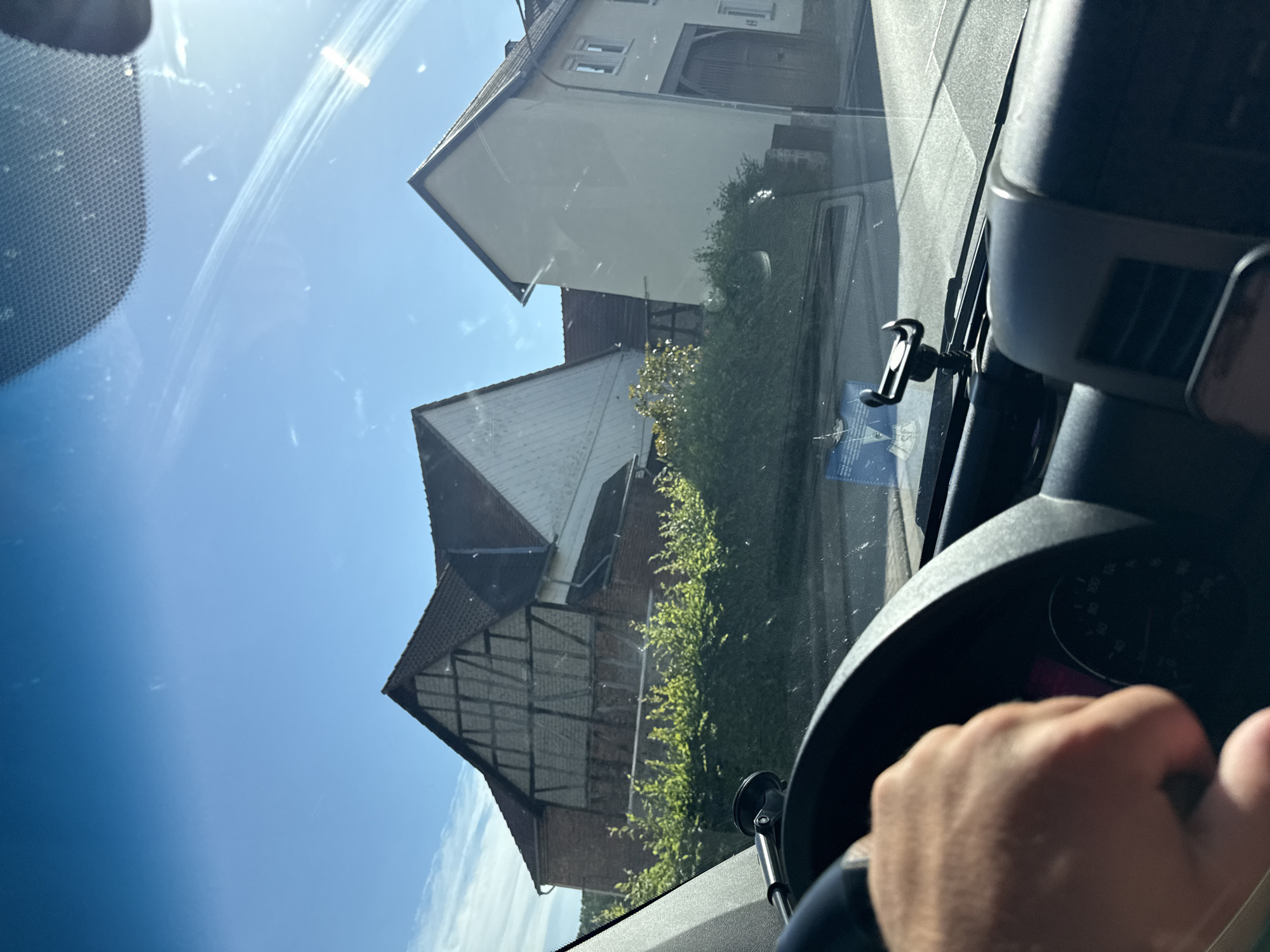 This media file is a first-person perspective shot from inside a vehicle, looking forward through the windshield. The image is taken on a clear, sunny day, suggesting it is daytime, likely mid-morning or afternoon given the bright sunlight and clear blue sky. The scene outside depicts a residential area with several houses visible. One house has a light-colored facade and a dark tiled roof, with visible window frames. Another building appears to be older, with a half-timbered upper section and a red brick lower section. A row of green bushes runs along the side of one of the buildings.
Inside the vehicle, a hand is visible gripping the steering wheel, indicating the person is driving. A black smartphone mount is attached to the windshield near the center of the frame. A portion of the car's dashboard, including the steering wheel and part of the instrument cluster, is also visible on the right side. The instrument cluster shows a dial with markings and numbers, suggesting it is a speedometer, with a portion of a digital display that appears to be pink.
The view through the windshield includes some smudges and reflections, characteristic of a car window. The overall mood of the image is bright and sunny, suggesting a pleasant day for driving. The location is identified as Volpertshausen, Germany. There is a small white sticker with blue text visible on the lower left side of the windshield, but the text is not legible.