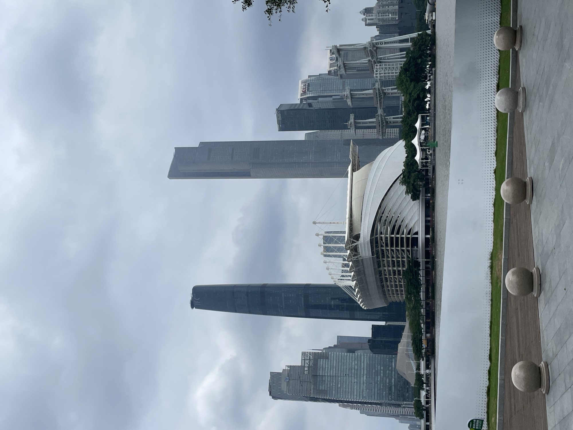 The image captures a skyline view in Yuexiu District, China, dominated by tall skyscrapers under a cloudy sky. Notable structures include the Canton Tower (Guangzhou TV Tower), and the Guangzhou Opera House, characterized by its modern, angular design. A paved walkway with spherical decorative elements runs along the foreground, adjacent to a narrow strip of green vegetation. The overall ambiance suggests an urban environment with a mix of architectural styles.