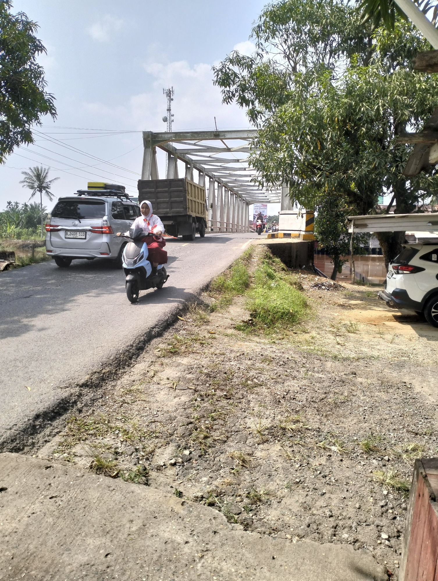In Mandala Jaya, Indonesia, a paved road runs parallel to a canal, leading towards a bridge. On the road, a woman wearing a white hijab and a red skirt rides a light blue scooter. Behind her, a silver SUV with a roof rack is stopped, and further ahead, a large truck is crossing the bridge. The bridge itself is an elevated structure with white support columns and a metal framework. Lush green trees with dense foliage are visible on the right side of the image, and on the left, palm trees and more trees are present. The ground is a mix of dirt, gravel, and sparse dry grass, with some patches of green grass near the road. The sky is clear and blue with scattered white clouds.