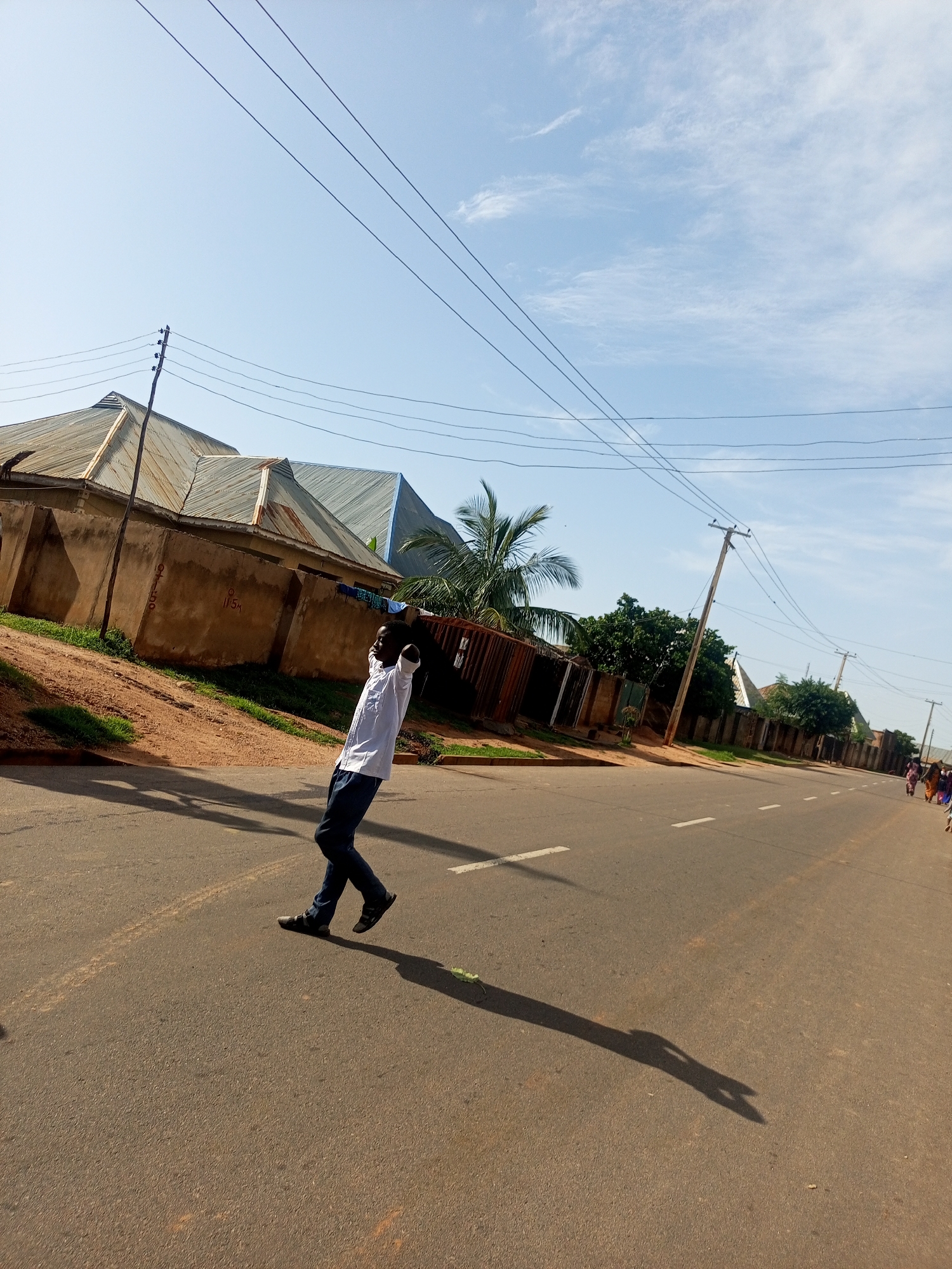 A young man in a white shirt and dark pants is walking across a paved road in Tempure, Nigeria. He is looking upwards with his arms raised behind his head. The road has white lane markings. In the background, there are buildings with corrugated metal roofs and a concrete wall. Several utility poles with wires are visible, along with some trees and a palm tree. The sky is blue with scattered clouds. A shadow of the man is cast on the road. In the far right background, a group of people can be seen walking.