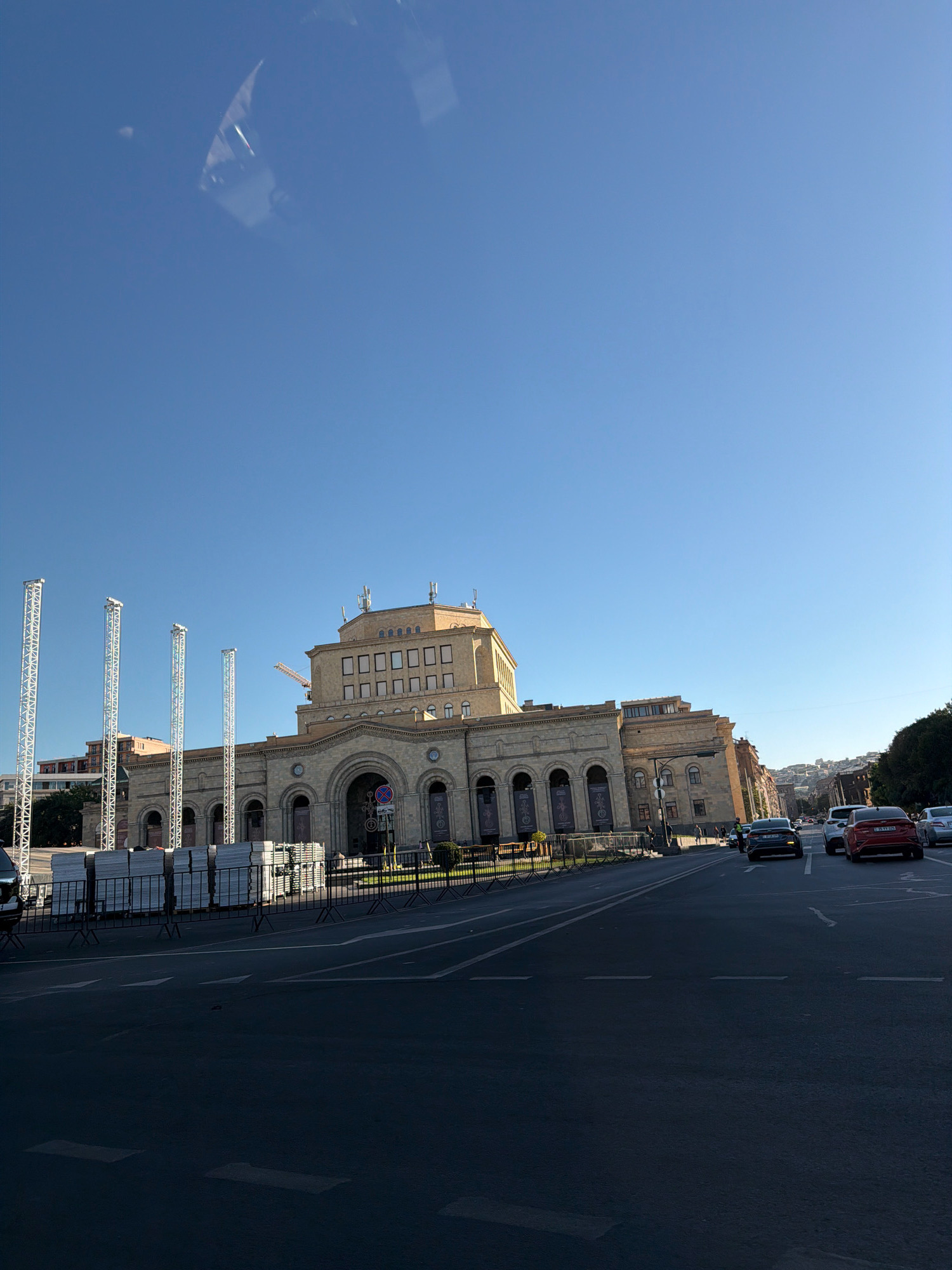 The image shows a wide, paved street in Yerevan, Armenia, with a large, tan-colored building occupying the left side of the frame. The building appears to be a public institution, possibly a theater or museum, with a grand arched entrance and multiple windows. In front of the building, several tall, metallic truss structures are erected, along with stacks of equipment covered in white tarps. There is a construction crane visible in the background, suggesting ongoing work. Cars are driving on the street, and a few pedestrians can be seen on the sidewalk in front of the building. The sky is clear and blue, indicating a sunny day. The overall scene suggests a vibrant urban environment, potentially preparing for an event or undergoing renovation.