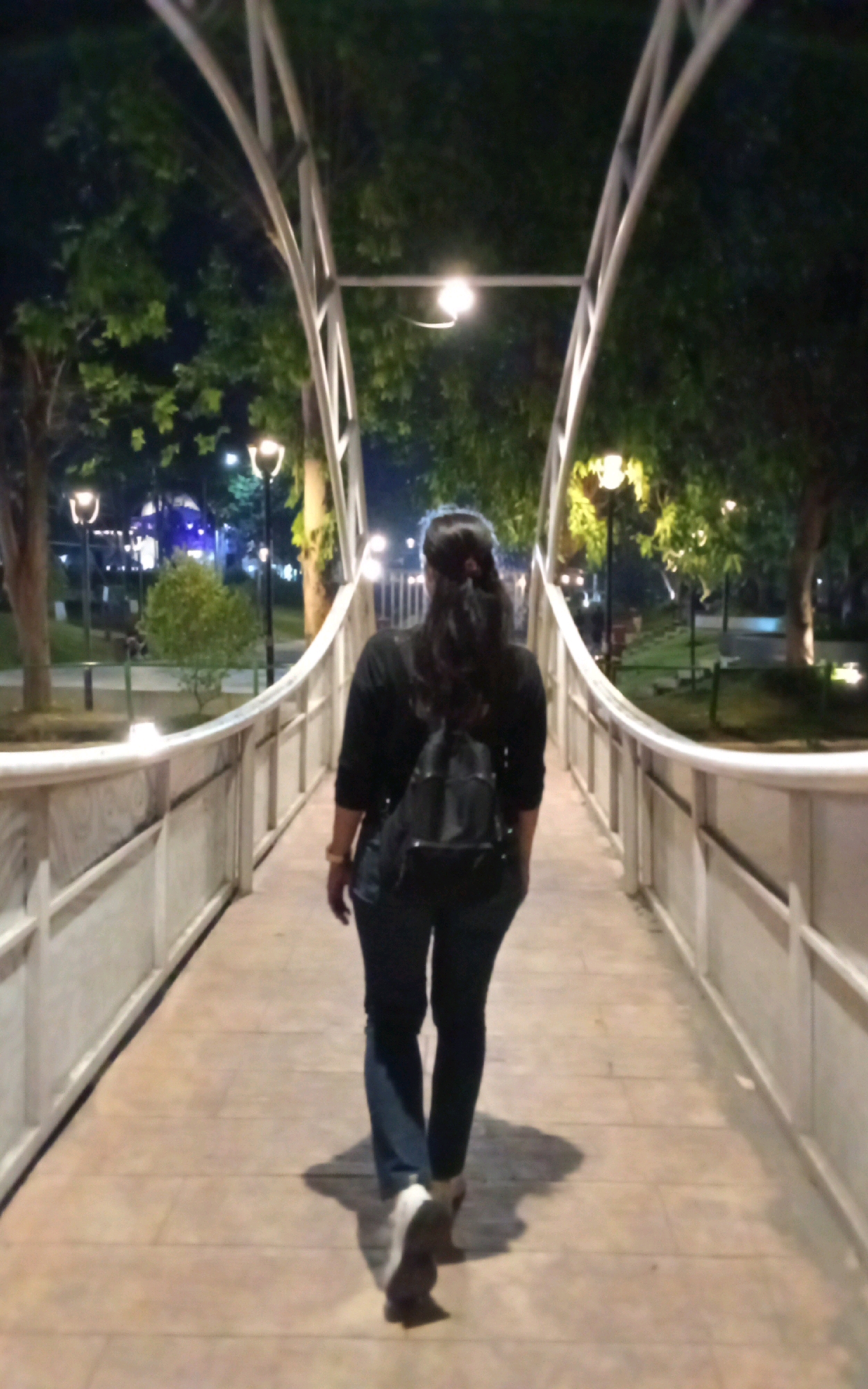A woman with dark hair pulled back is walking away from the camera on a pedestrian bridge at night. She is wearing a black long-sleeved shirt, dark blue jeans, and white and brown shoes. She is also carrying a black backpack. The bridge has white railings and is illuminated by streetlights. Trees and foliage are visible on either side of the bridge, and other lights are visible in the background. The scene appears to be in a park or public space within the city of Ngasem, Indonesia.